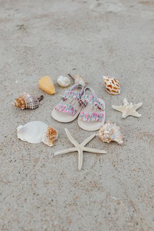 Colorful girls sandals surrounded by seashells and starfish on sandy beach.