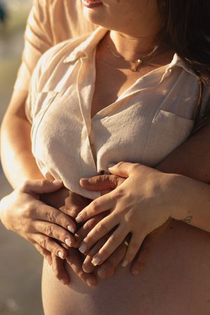 Pregnant woman in sheer blouse with partner's hands tenderly cradling her baby bump in warm golden sunlight.