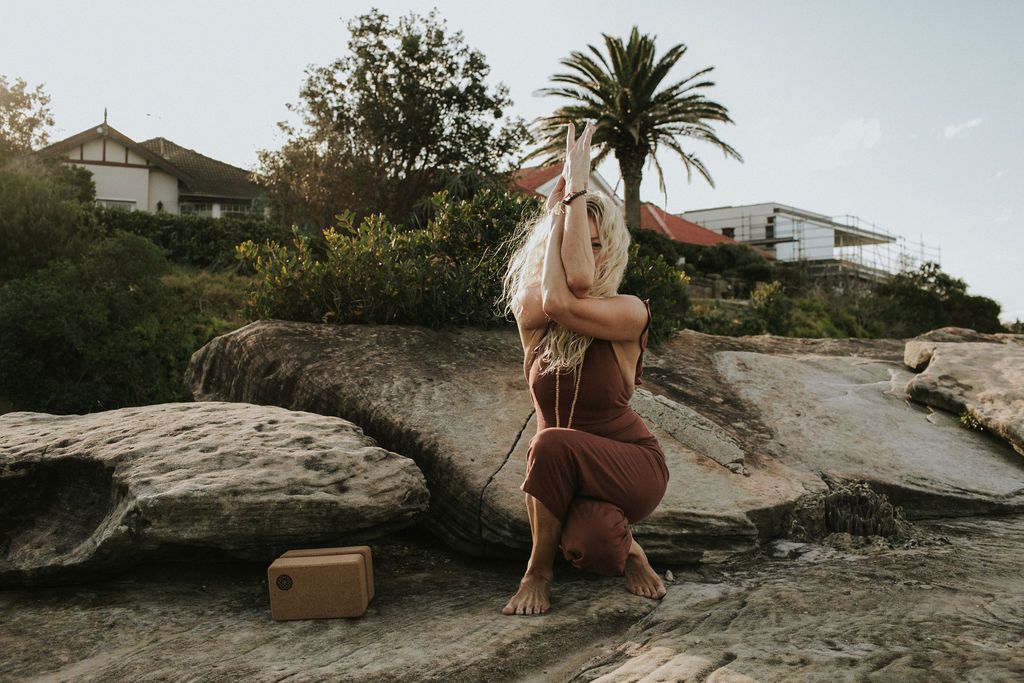 Woman practicing yoga on coastal rocks near a palm tree and white house, wearing a brown outfit in golden hour light.