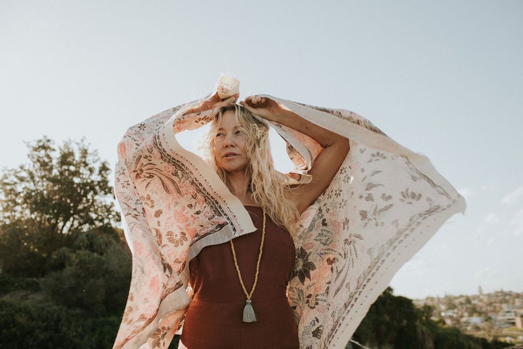 Boho woman with blonde hair holding floral scarf overhead outdoors in golden hour light, wearing rust dress and gold necklace.
