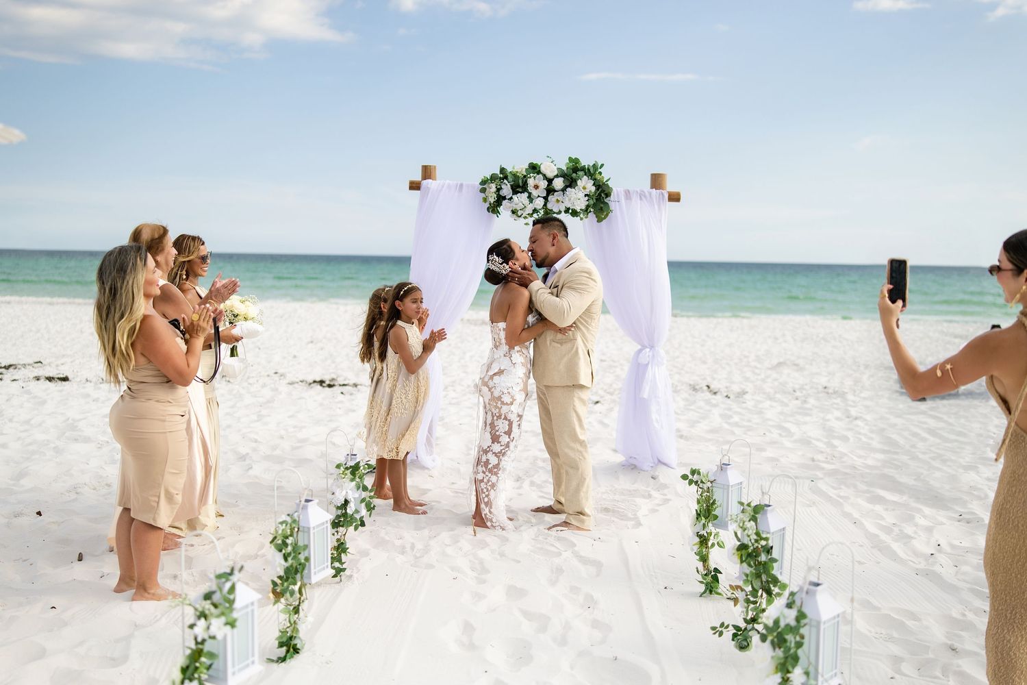 Couple shares first kiss at romantic beach wedding ceremony with white arch, floral decor, and bridal party on white sand.