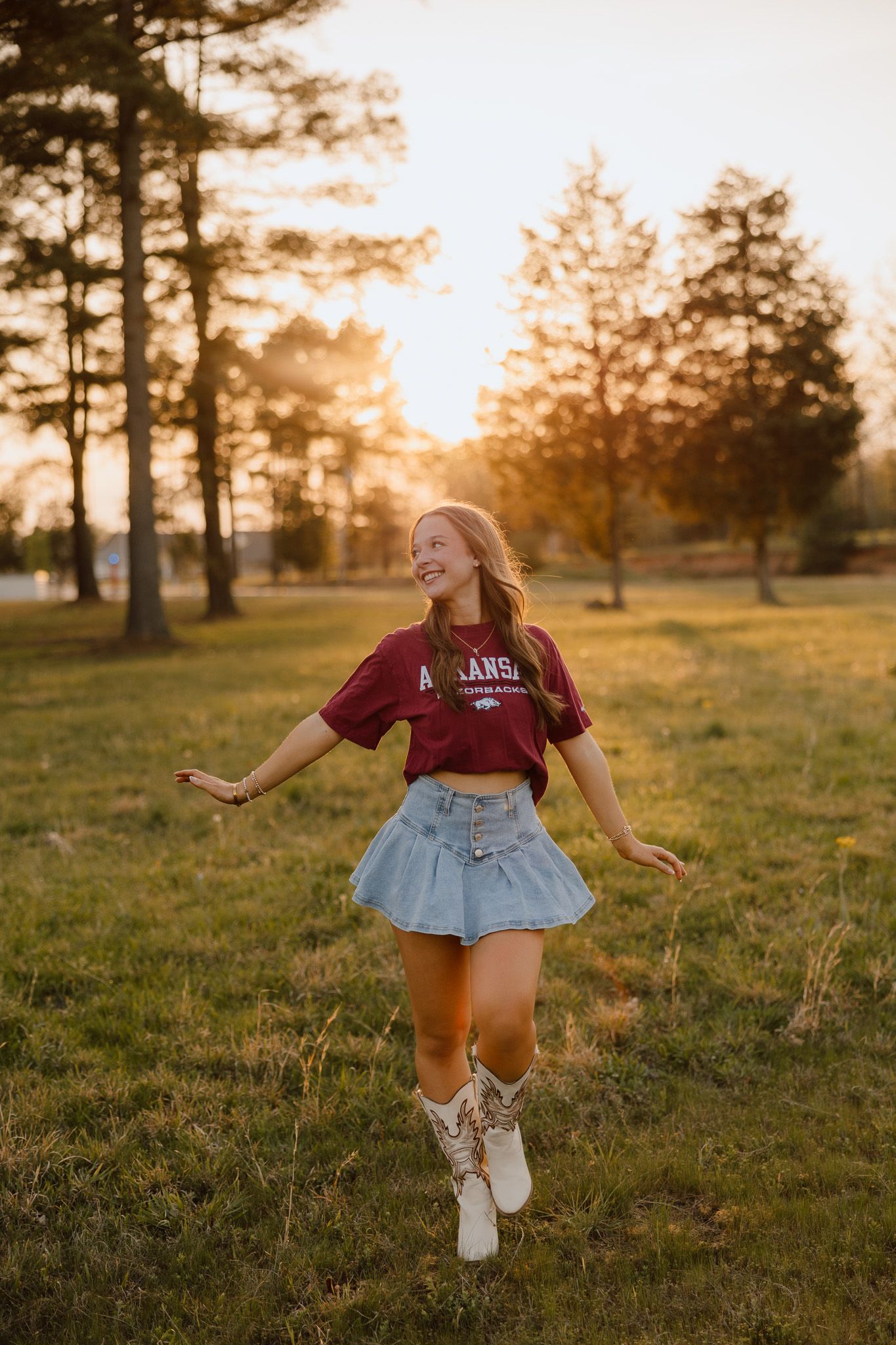 Young woman in maroon Arkansas shirt, denim skirt, and cowboy boots dancing in a sunlit park at golden hour.