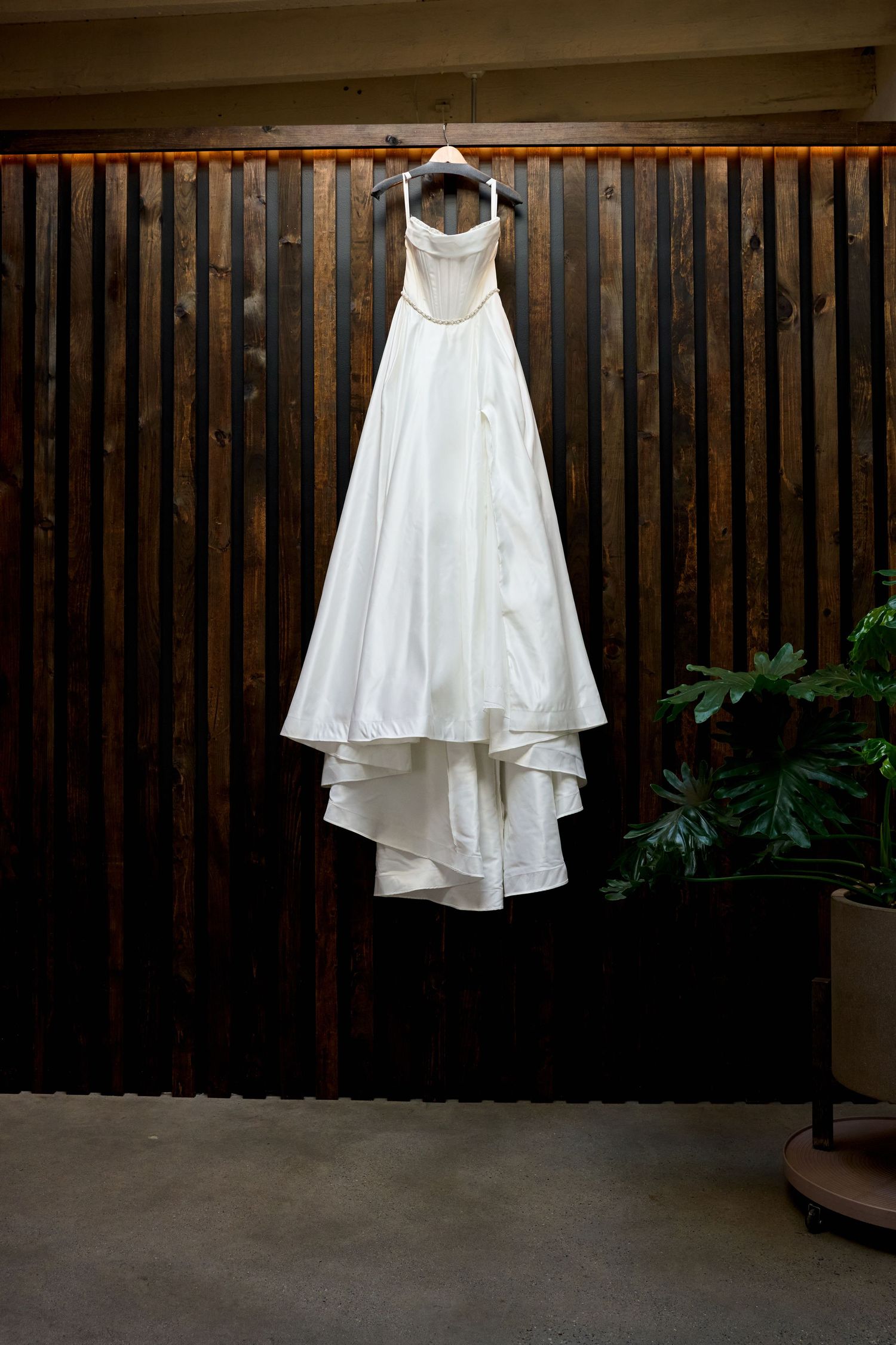 White satin A-line wedding dress with thin straps hanging on wooden slat wall beside tropical plant.