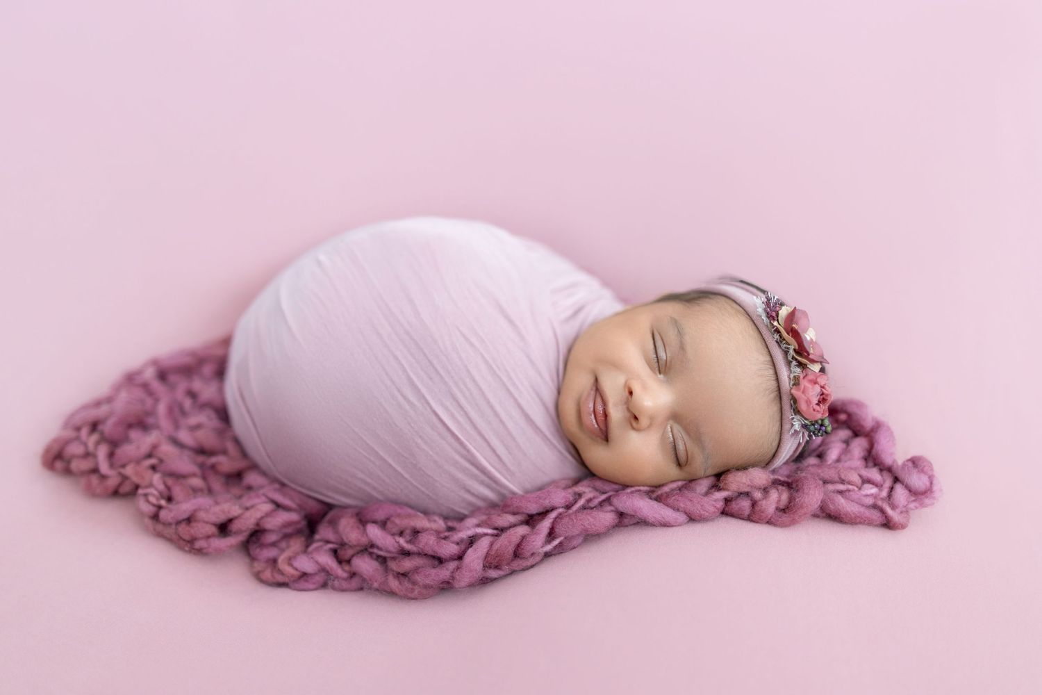 Newborn baby girl swaddled in lavender wrap, sleeping on pink chunky knit blanket with floral headband.