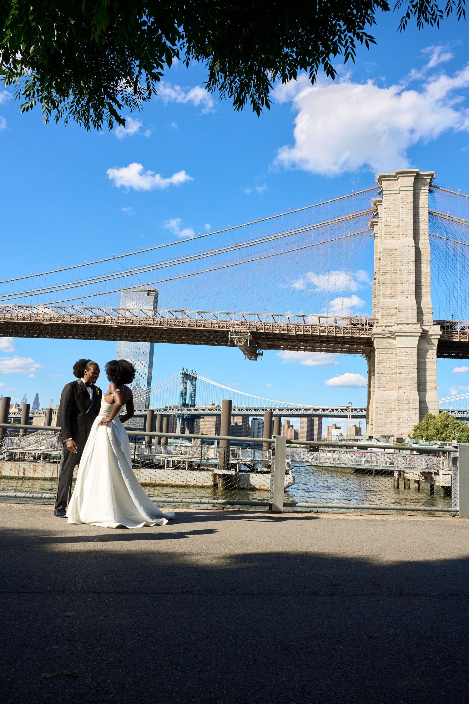 Newlywed couple poses near Brooklyn Bridge waterfront on sunny day with blue sky and Manhattan skyline backdrop.