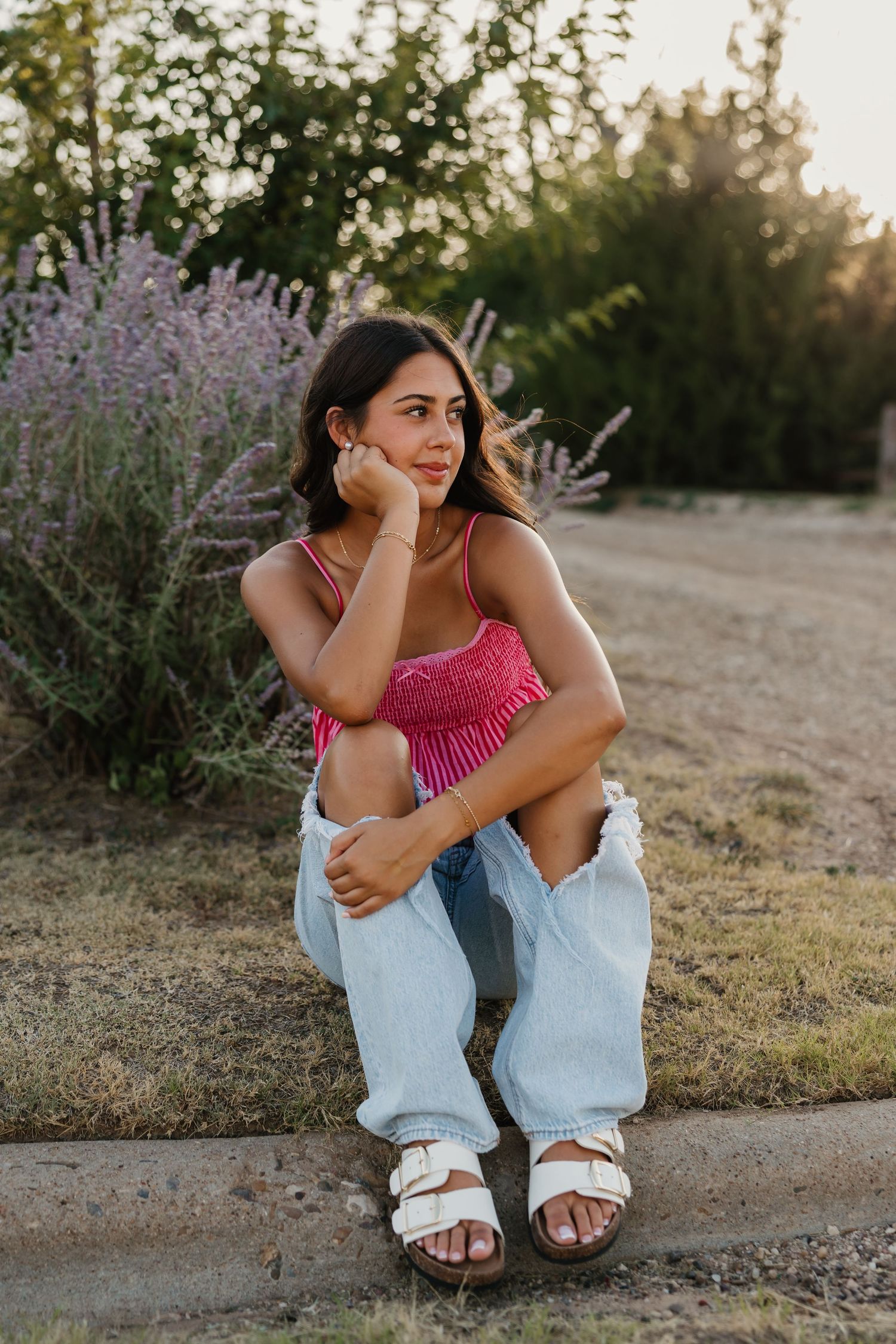 A brunette girl wearing pink top and ripped blue jeans poses for senior portraits in front of purple flowers in Tulia, Texas