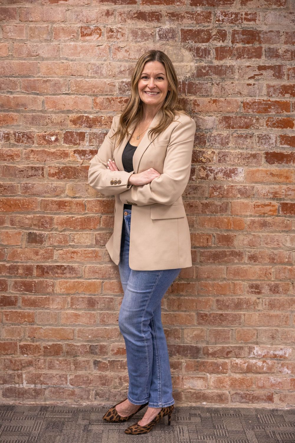 Woman in beige blazer and jeans smiling with arms crossed against a rustic red brick wall.