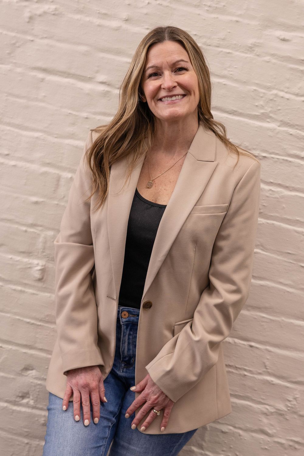 Smiling woman in beige blazer and jeans posing against white brick wall.