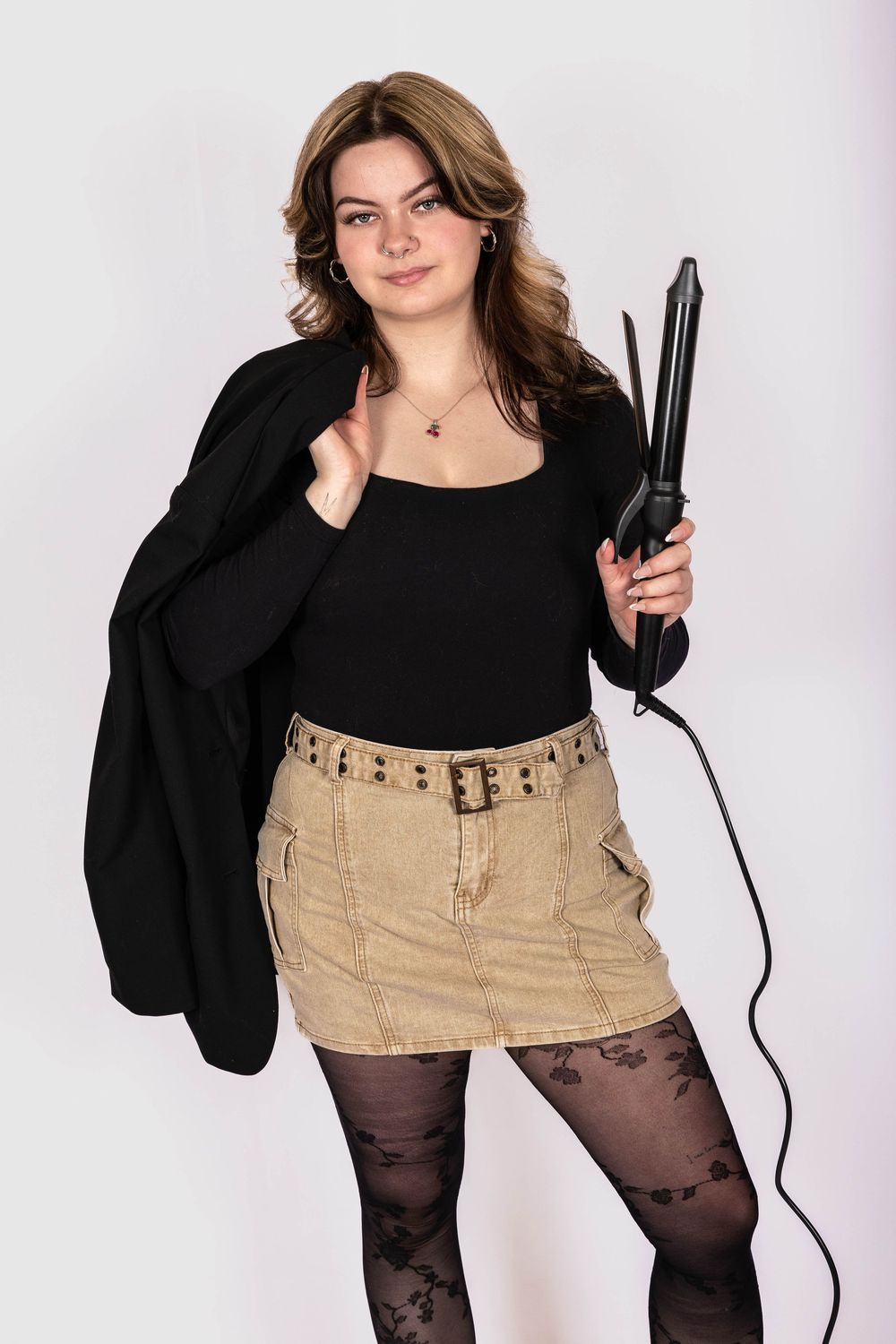 Young woman in black top, tan mini skirt, and patterned tights holds a curling iron and jacket on white background.