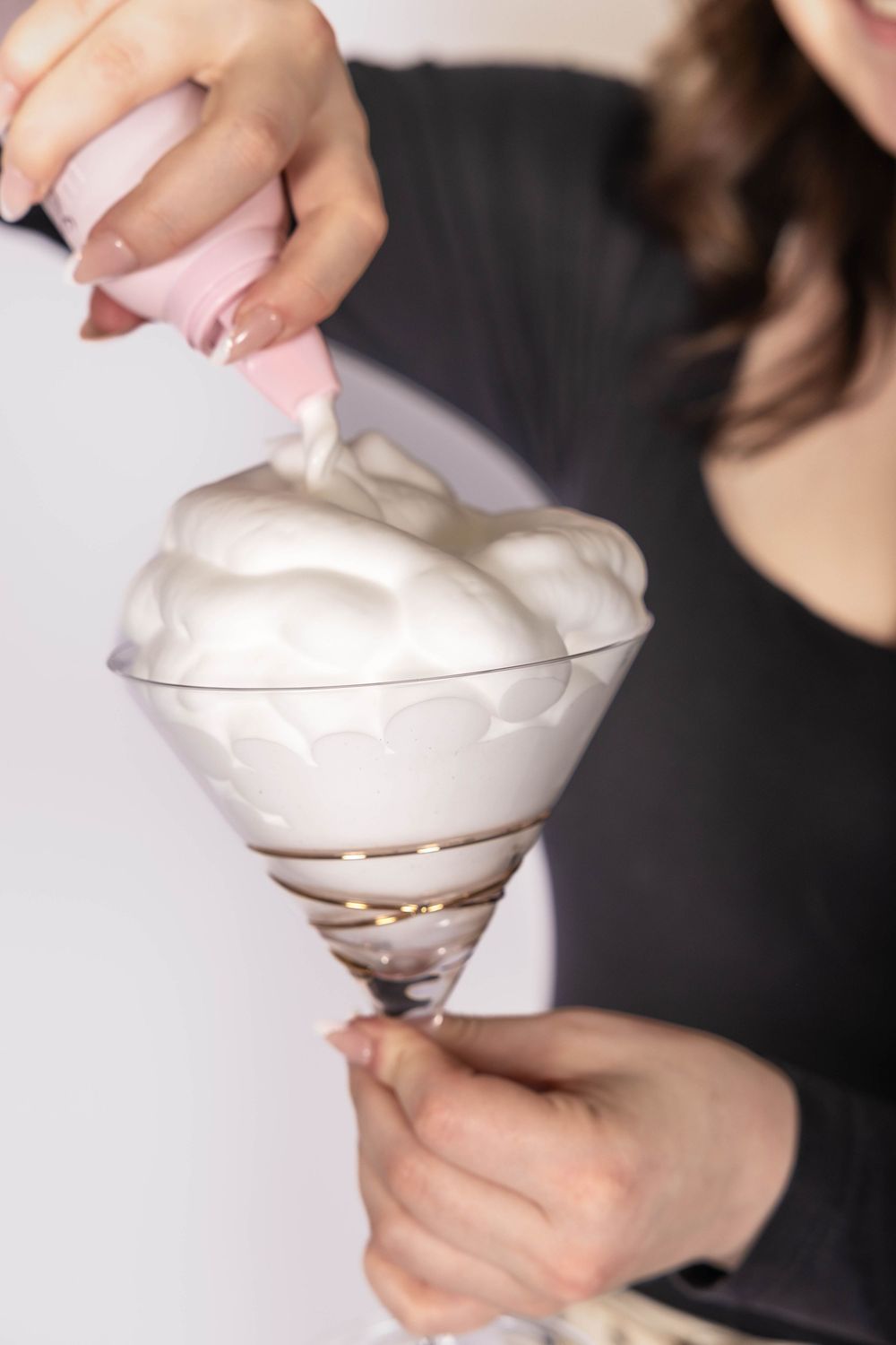 Woman in black top piping white whipped cream into a decorative glass dessert cup.