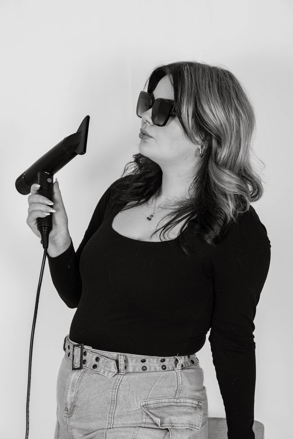 Black and white photo of a woman in sunglasses holding a hair dryer, wearing a black top and jeans.