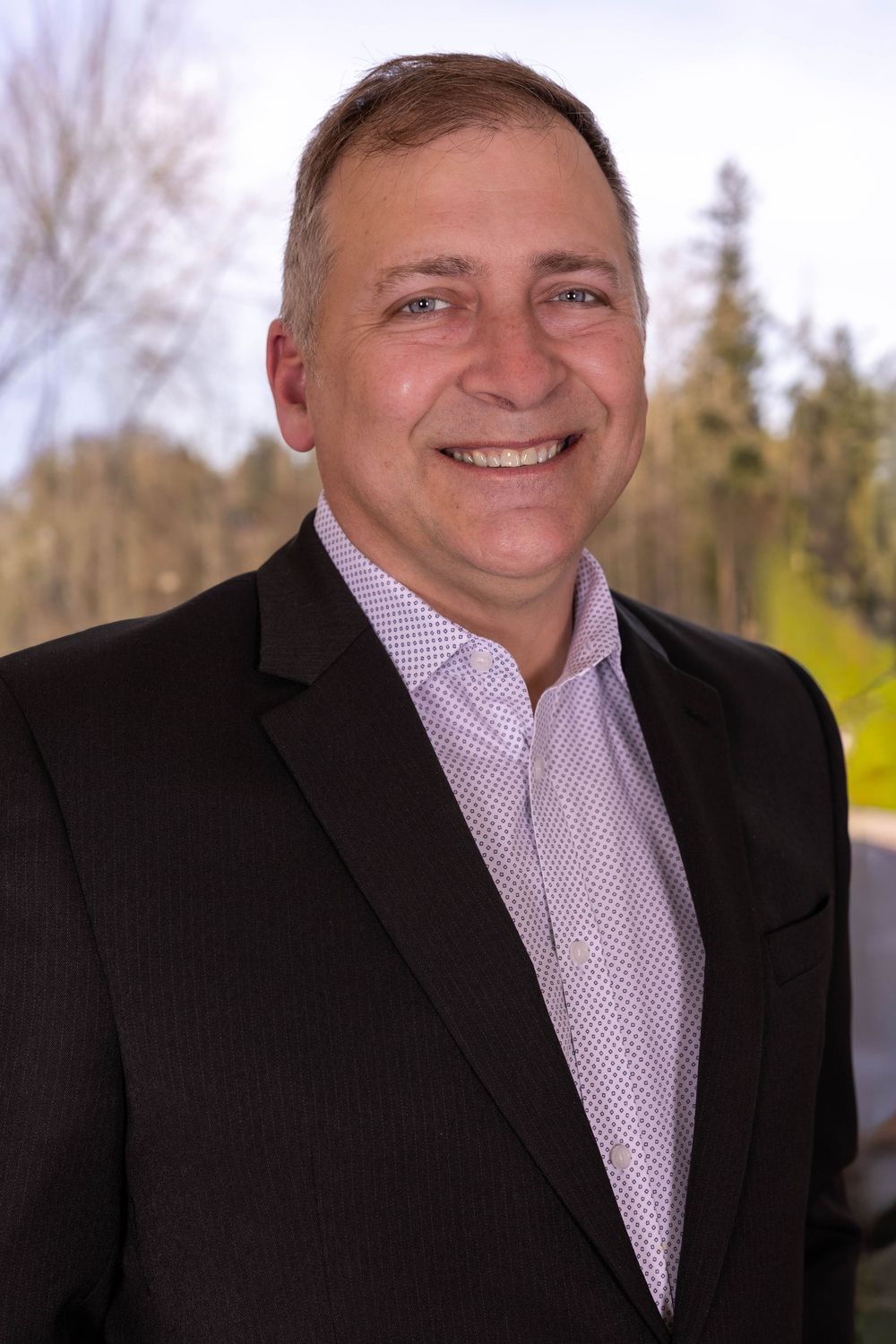 Professional man in dark blazer smiling outdoors with trees in background, ideal corporate headshot photo.