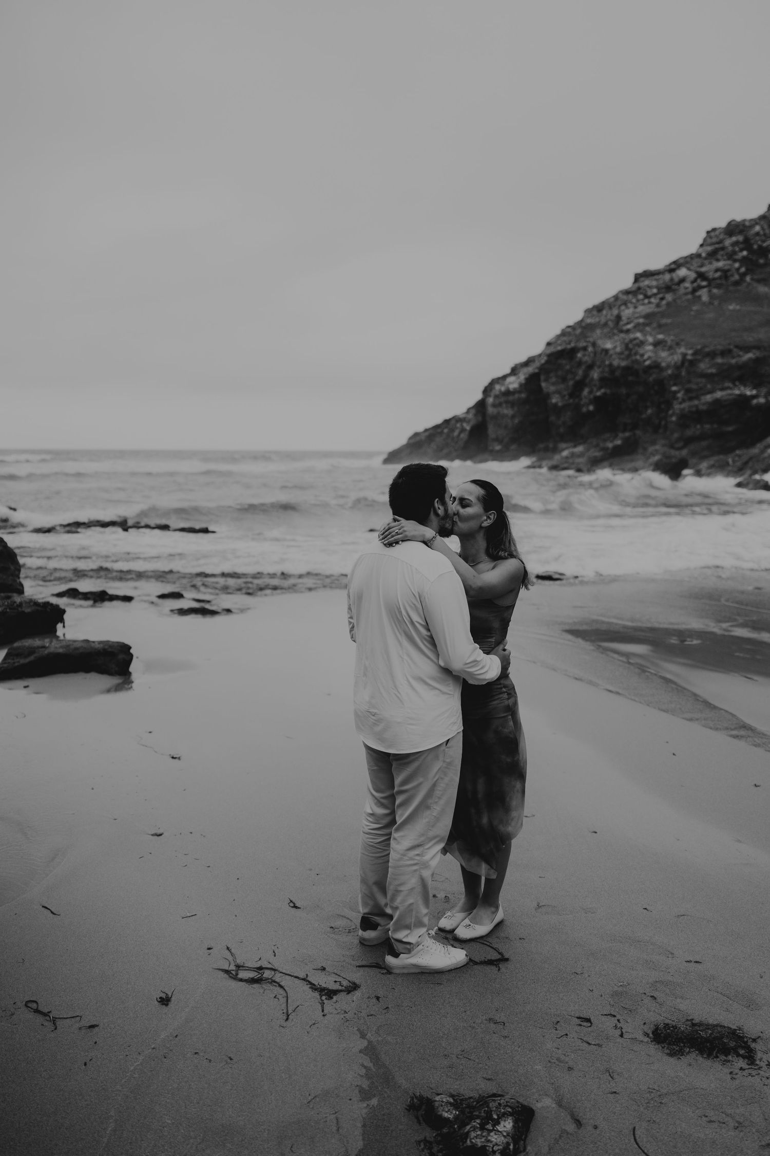 Black and white photo of a couple kissing on chapel porth beach with rocky cliffs and ocean waves in the background.