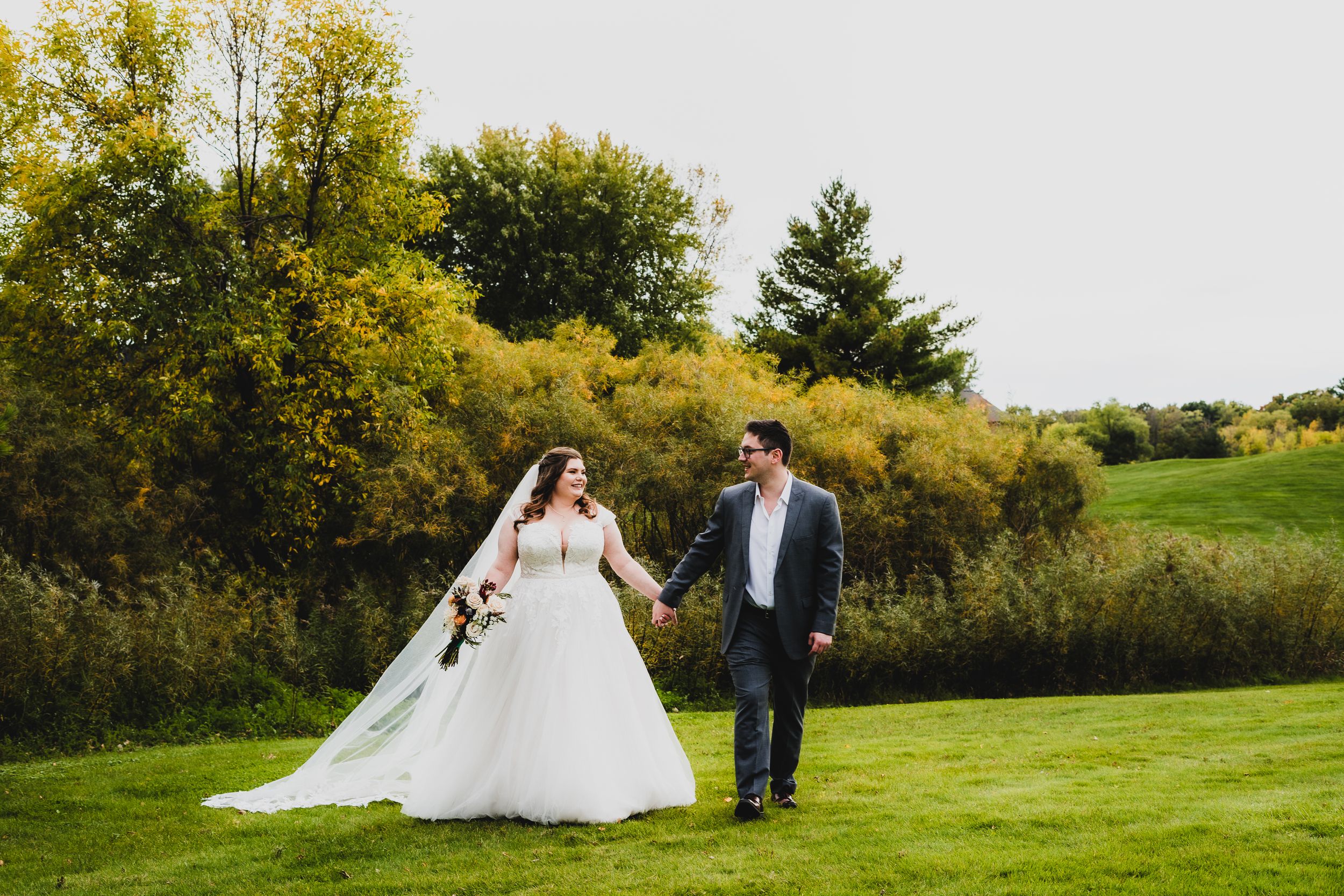 Bride and groom walking together on lush green lawn with autumn trees in background.