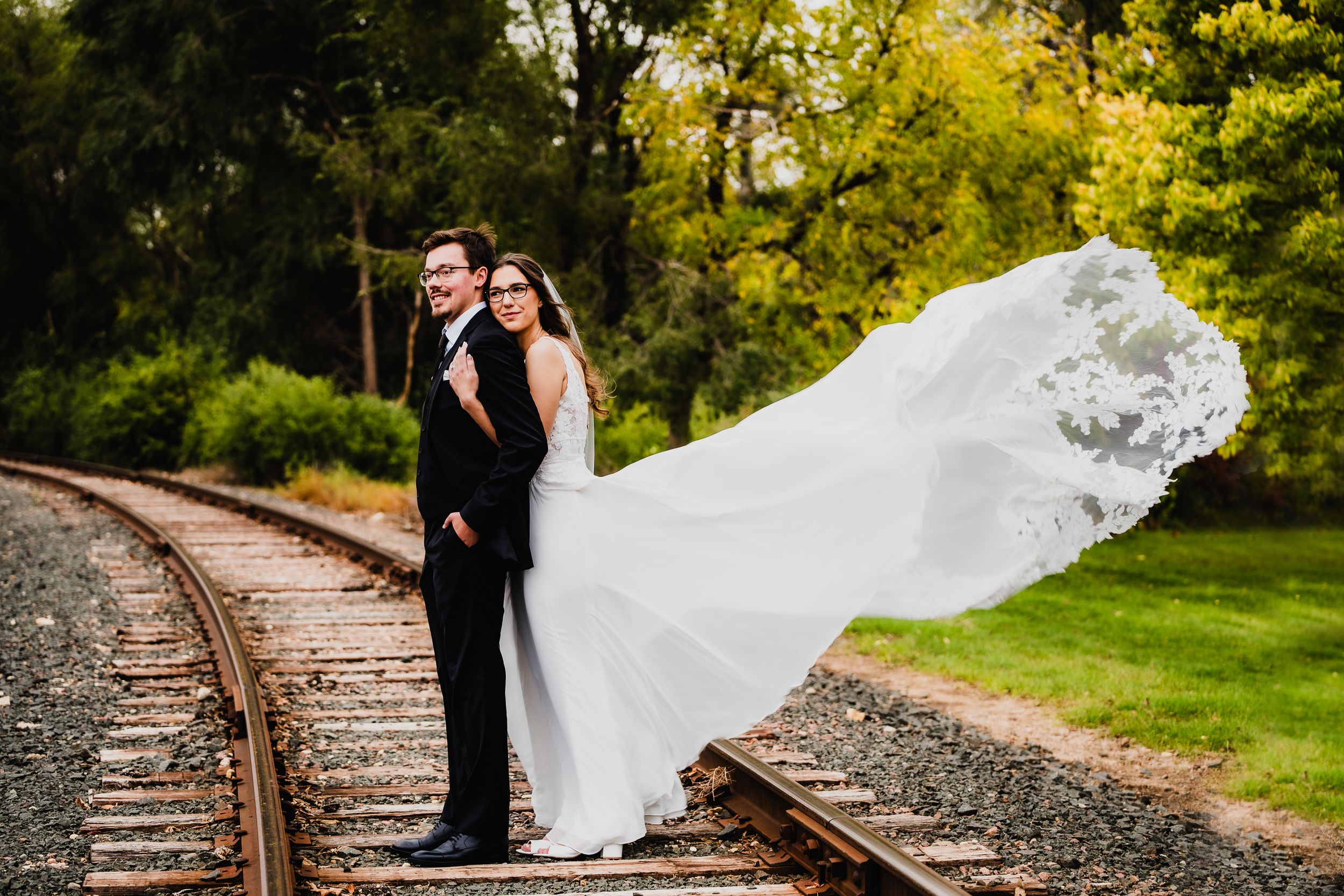 Wedding couple poses on railroad tracks with flowing veil in natural setting.