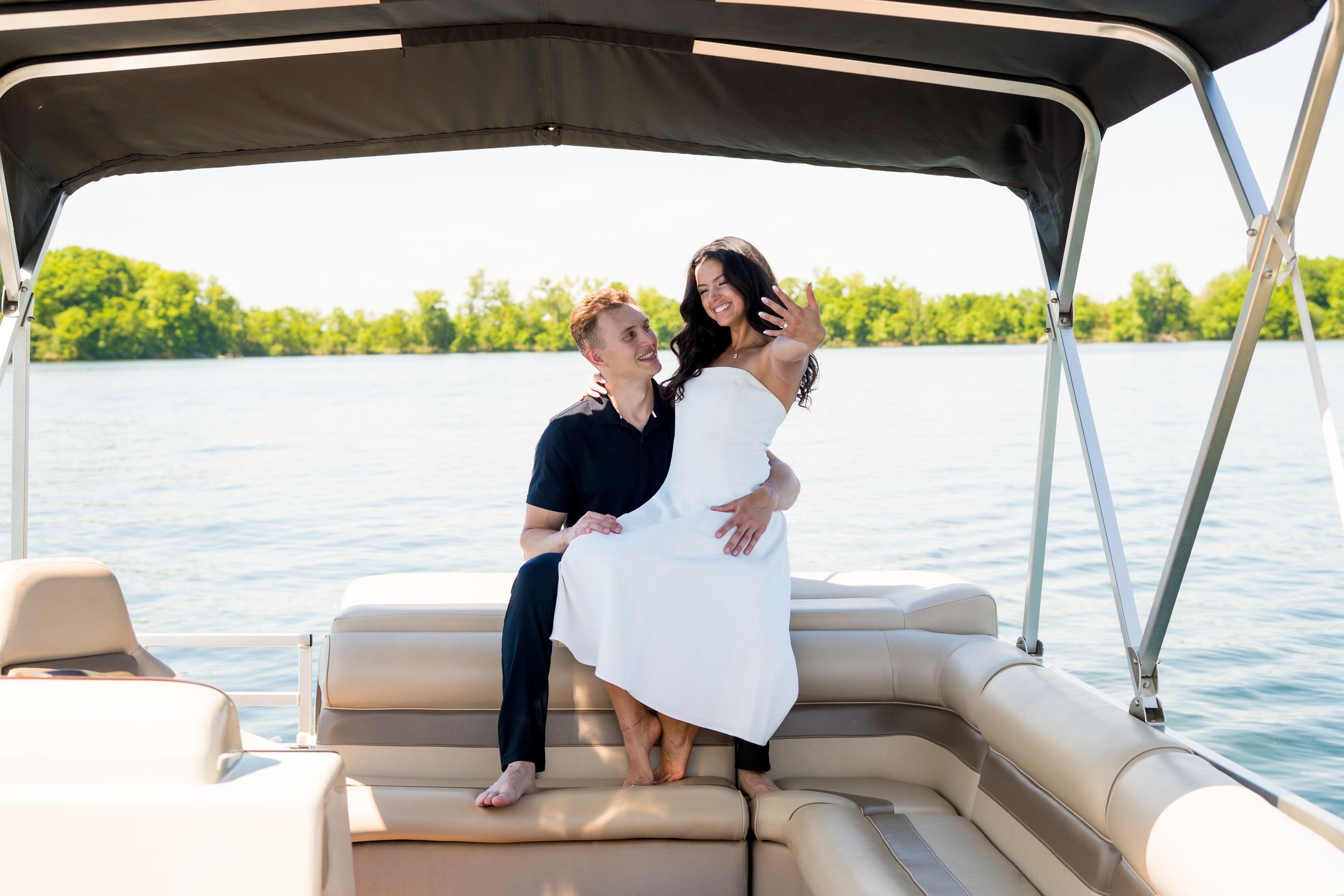 Couple embracing on a pontoon boat on a calm lake, woman in white dress, man in black outfit, surrounded by lush trees.