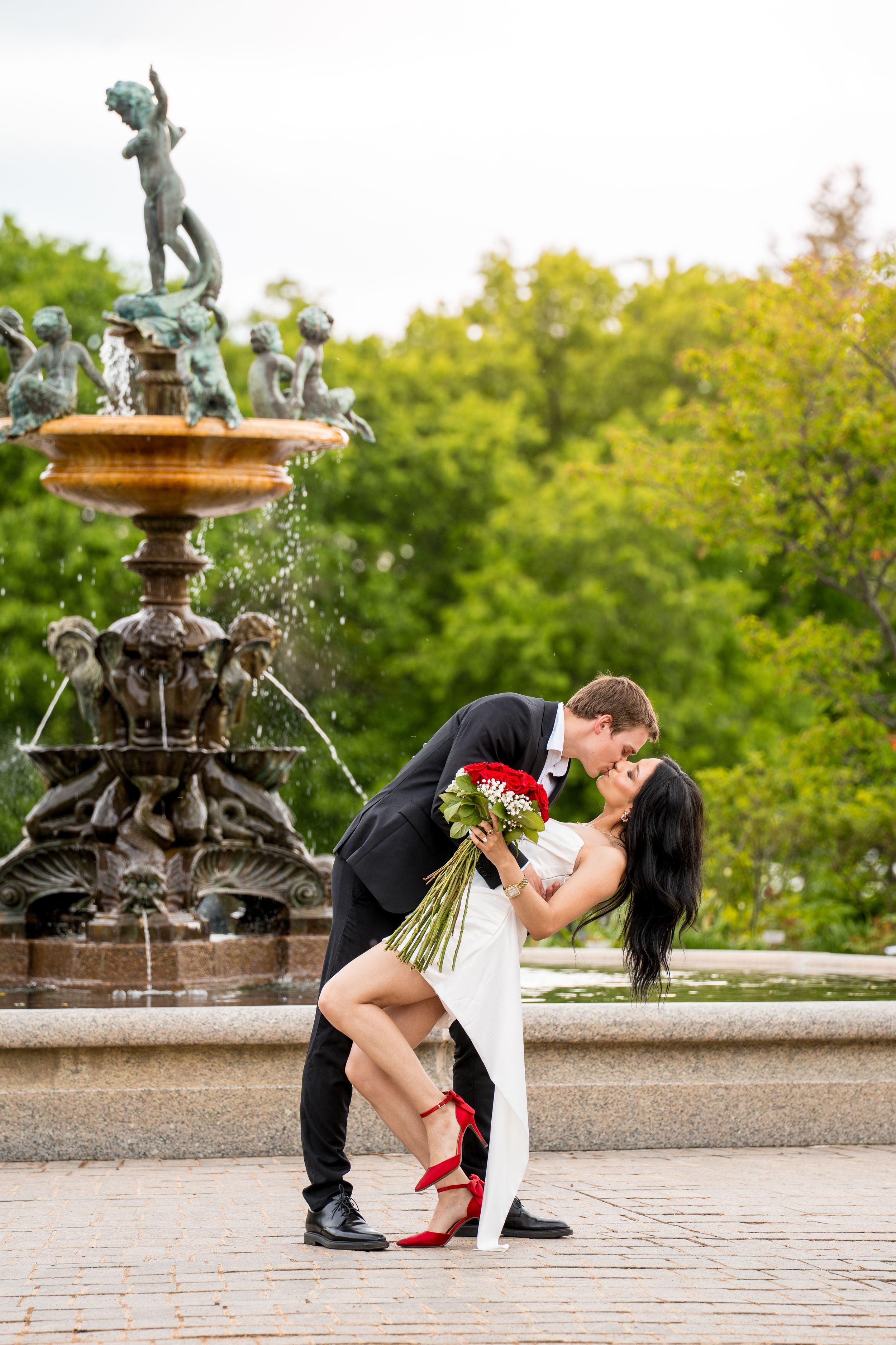 Couple sharing a romantic dip kiss in front of an ornate fountain, bride holding red roses in white dress and red heels.