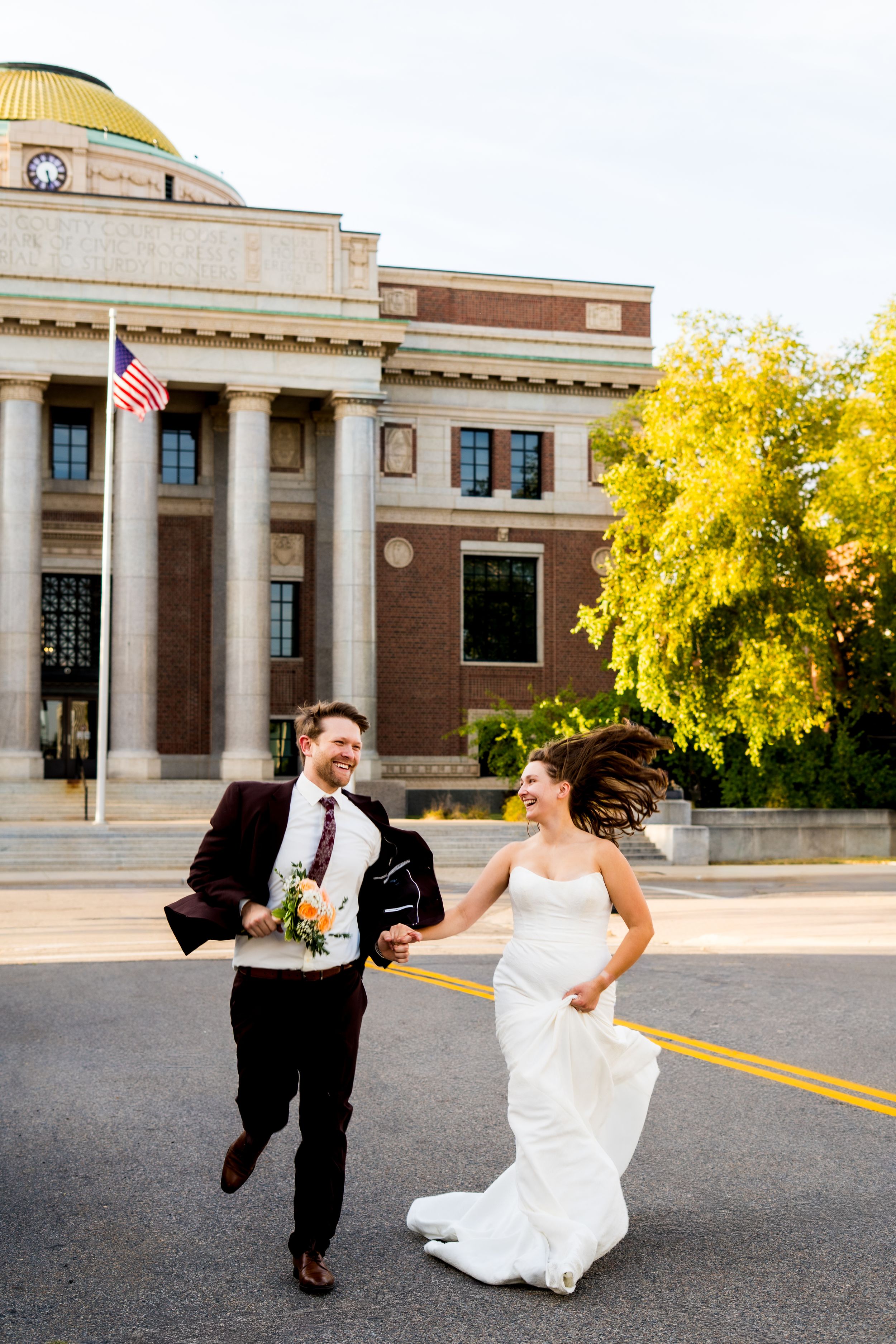 A joyful couple runs and laughs together on their wedding day in front of a grand building with columns and a golden dome.