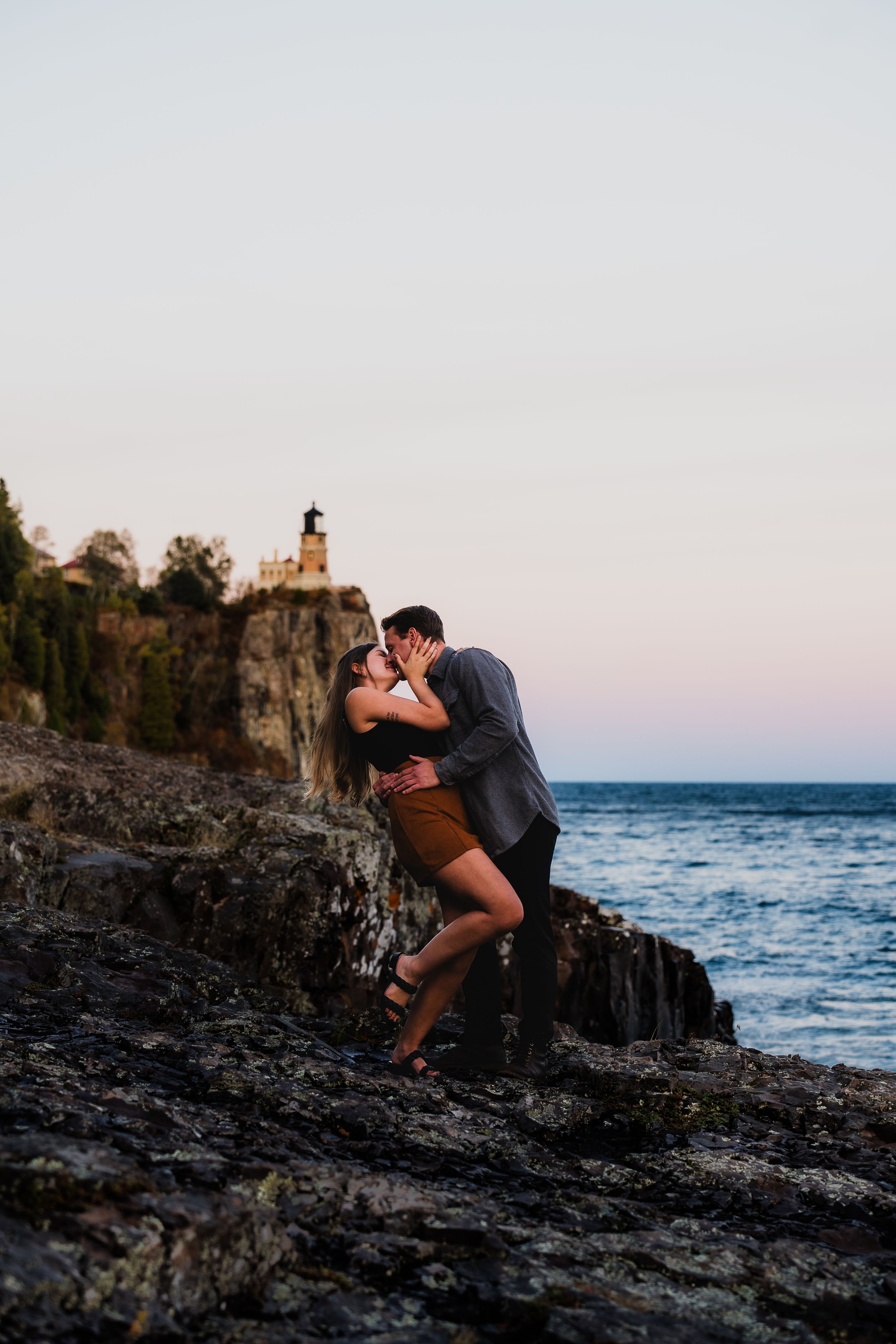 Couple sharing a romantic kiss on rocky shoreline at sunset with a lighthouse on cliffs in the background.