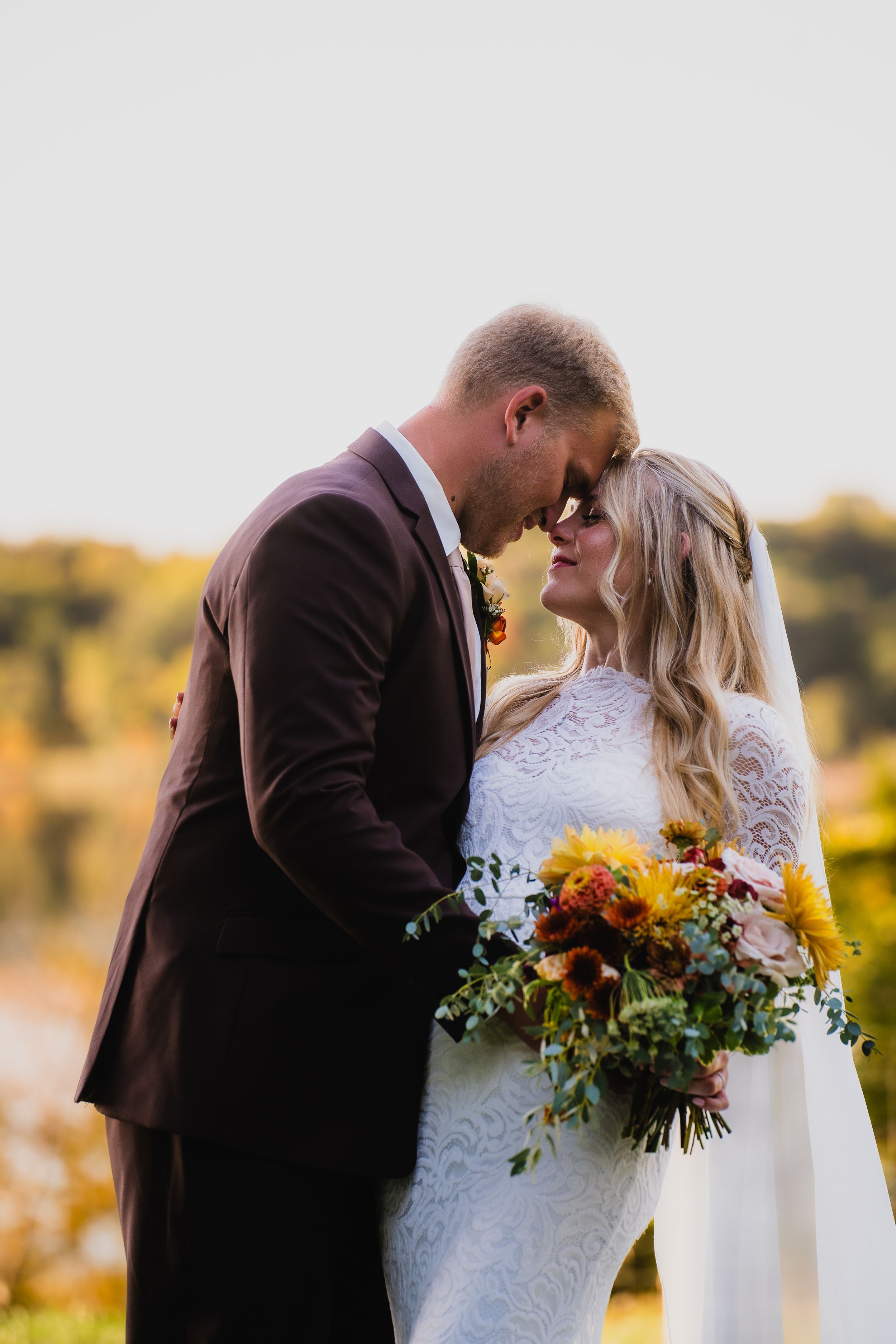 Newlywed couple shares intimate moment at sunset holding vibrant sunflower bouquet in rustic outdoor setting.