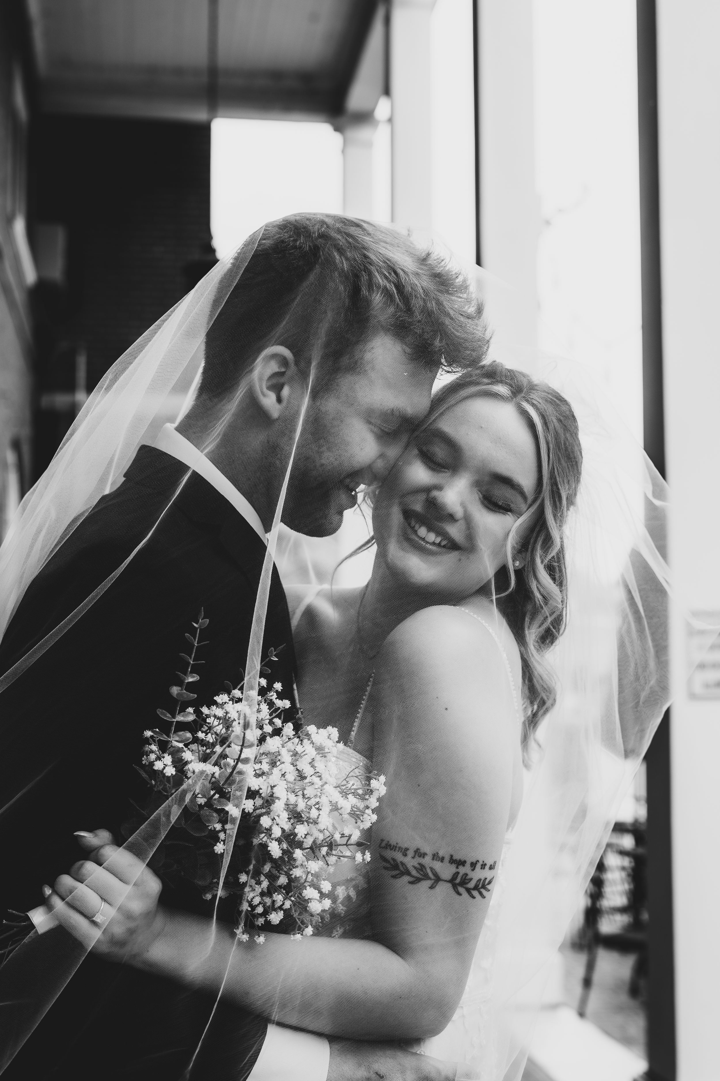A tender black and white wedding photo showing a joyful bride in white dress and groom with baby's breath bouquet.