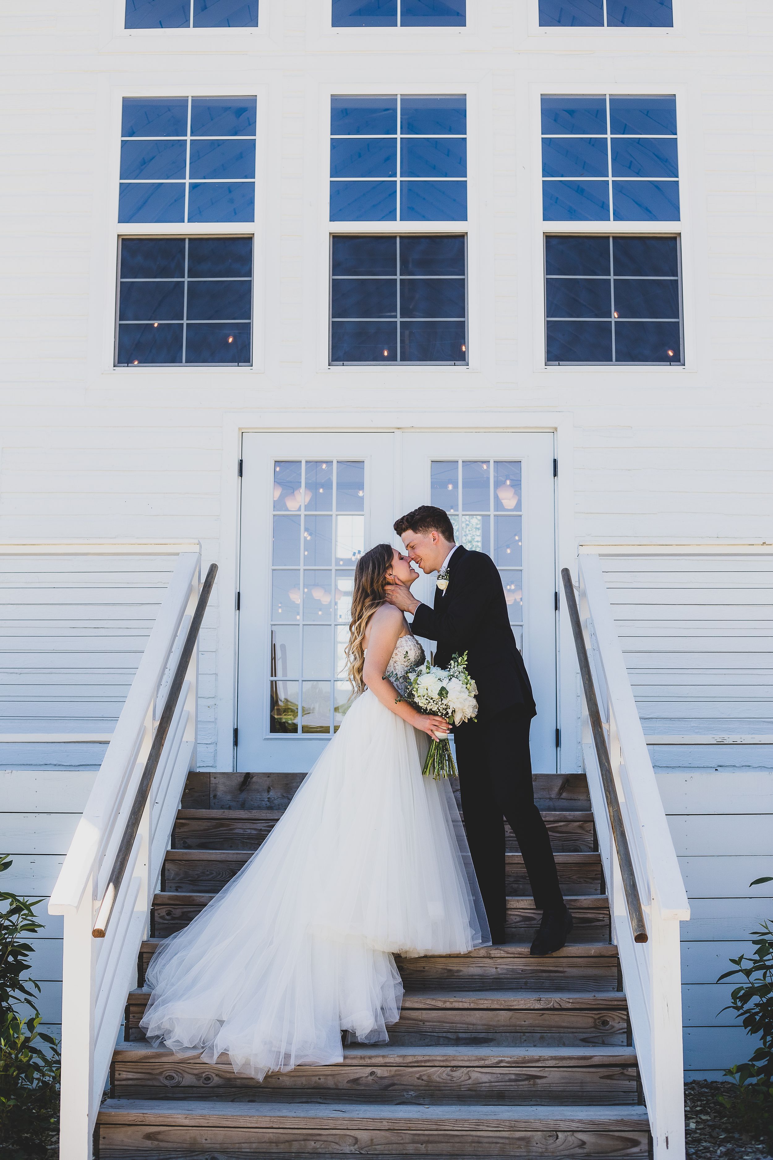 A wedding couple shares a tender moment on the steps of a white church building with symmetrical windows.