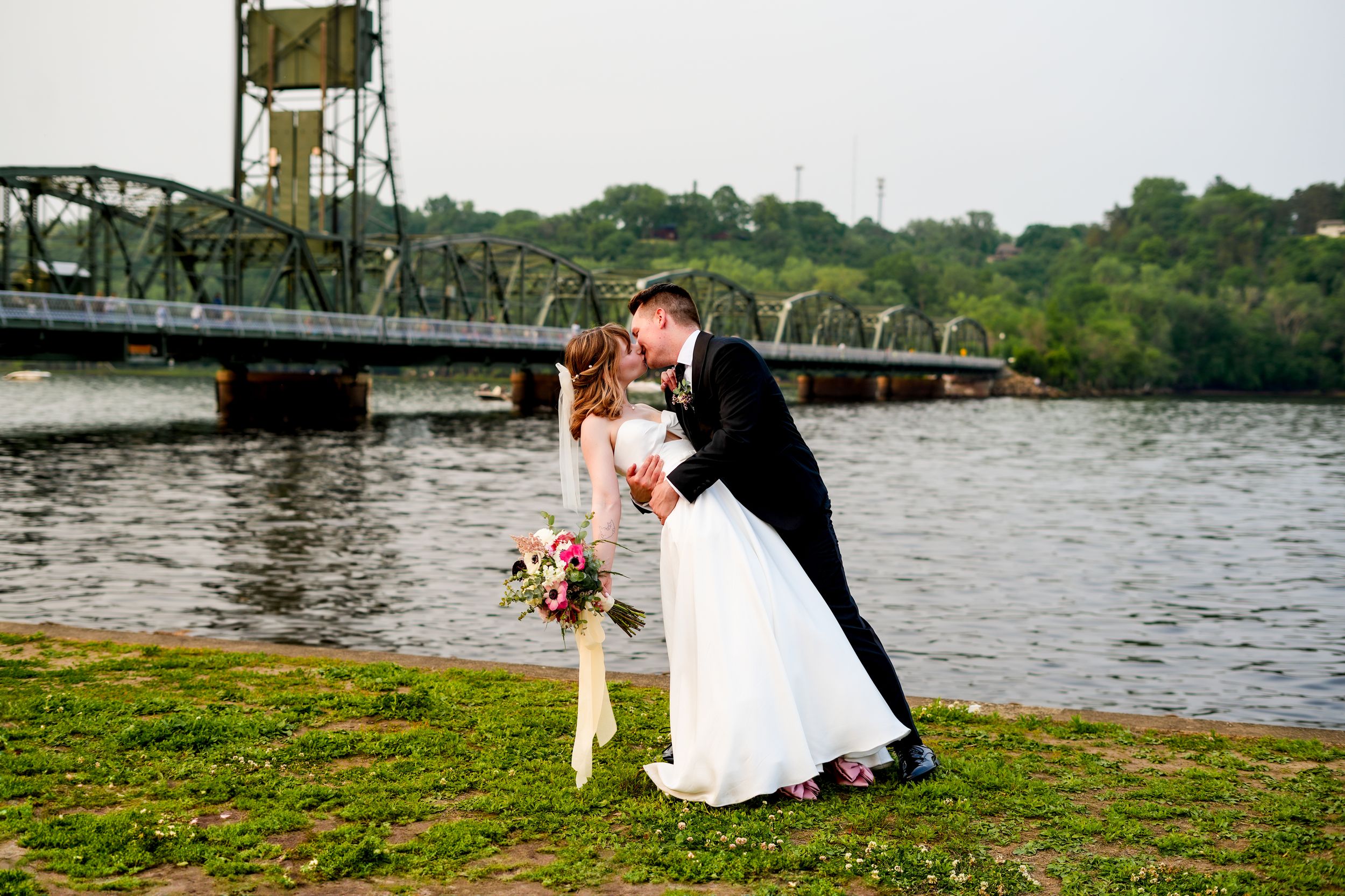 A couple shares a romantic moment by a river with an iron bridge in the background.
