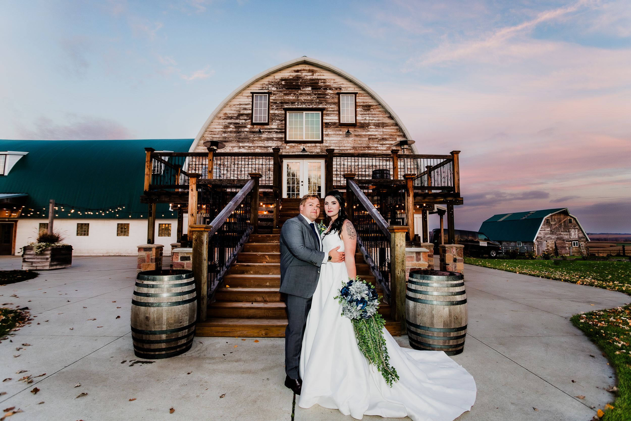 Elegant couple poses in front of rustic barn venue with wine barrels at sunset.