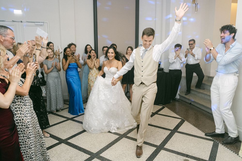 Bride and groom make grand entrance at wedding reception, guests cheering and clapping on dance floor.