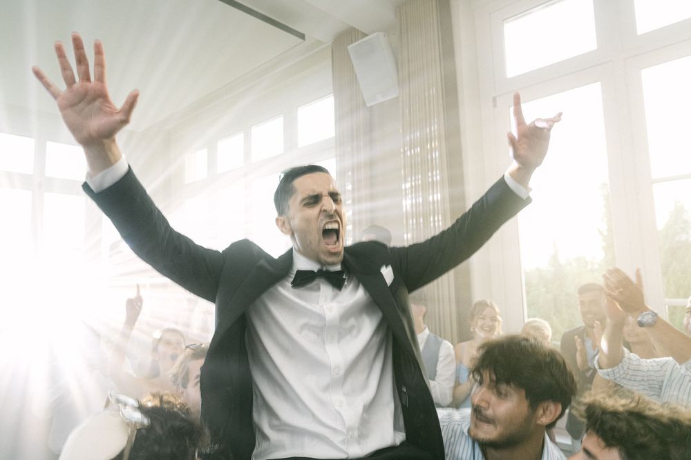 Excited groom in tuxedo raises arms triumphantly surrounded by cheering wedding guests in bright reception hall.