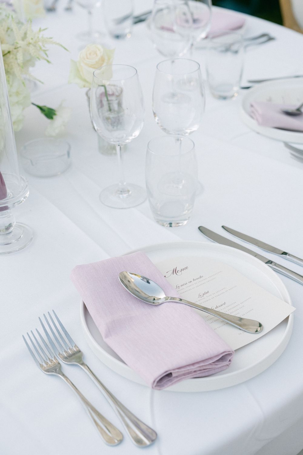 Elegant wedding table setting with white plates, lavender napkins, silver cutlery, crystal glasses, and white floral centerpiece.