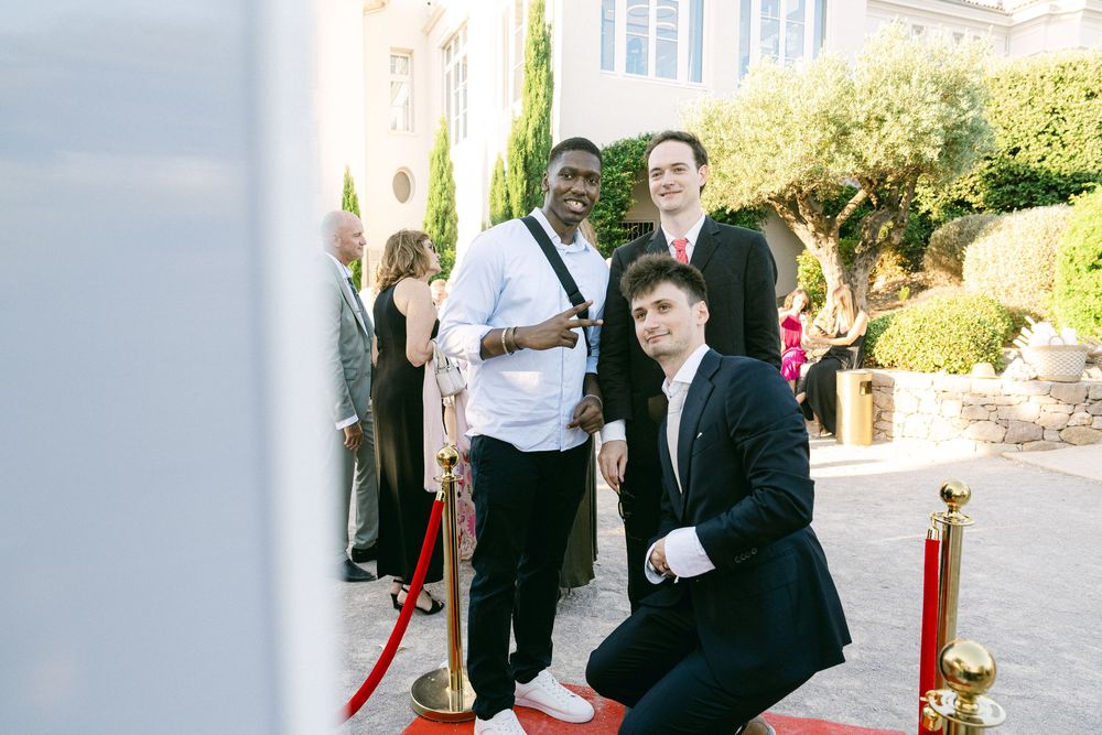 Three men posing on a red carpet at a formal outdoor event near a villa.