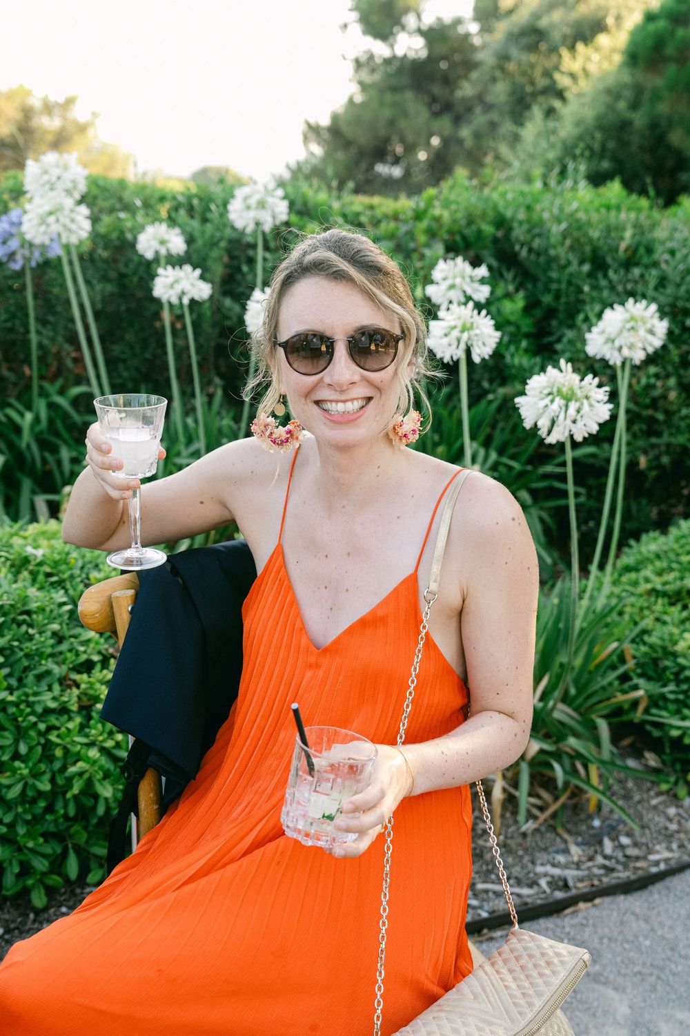 Smiling woman in orange dress holding drinks outdoors, wearing sunglasses near white agapanthus flowers.