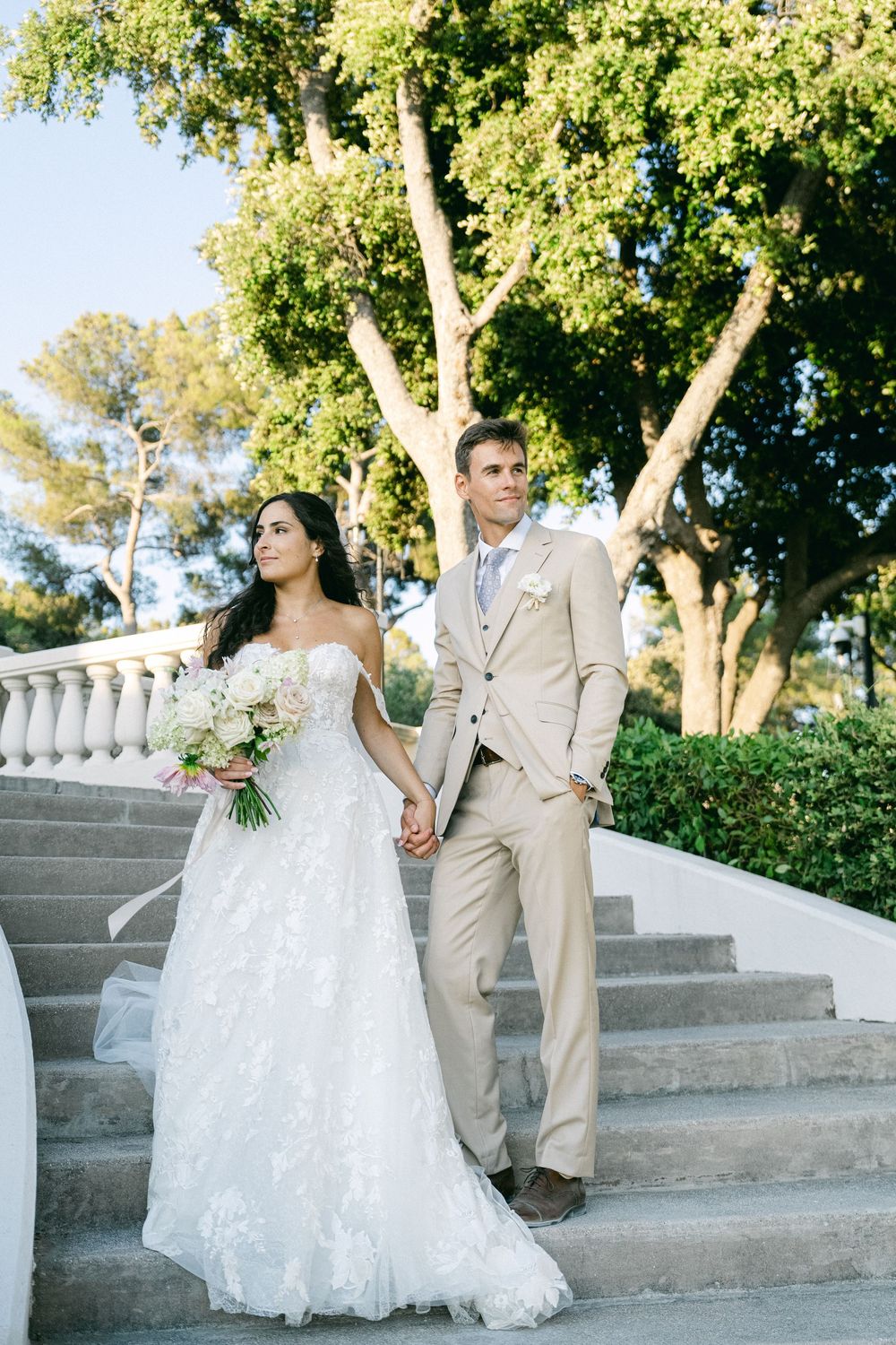 Bride in lace gown and groom in tan suit hold hands on outdoor stone steps surrounded by lush greenery.