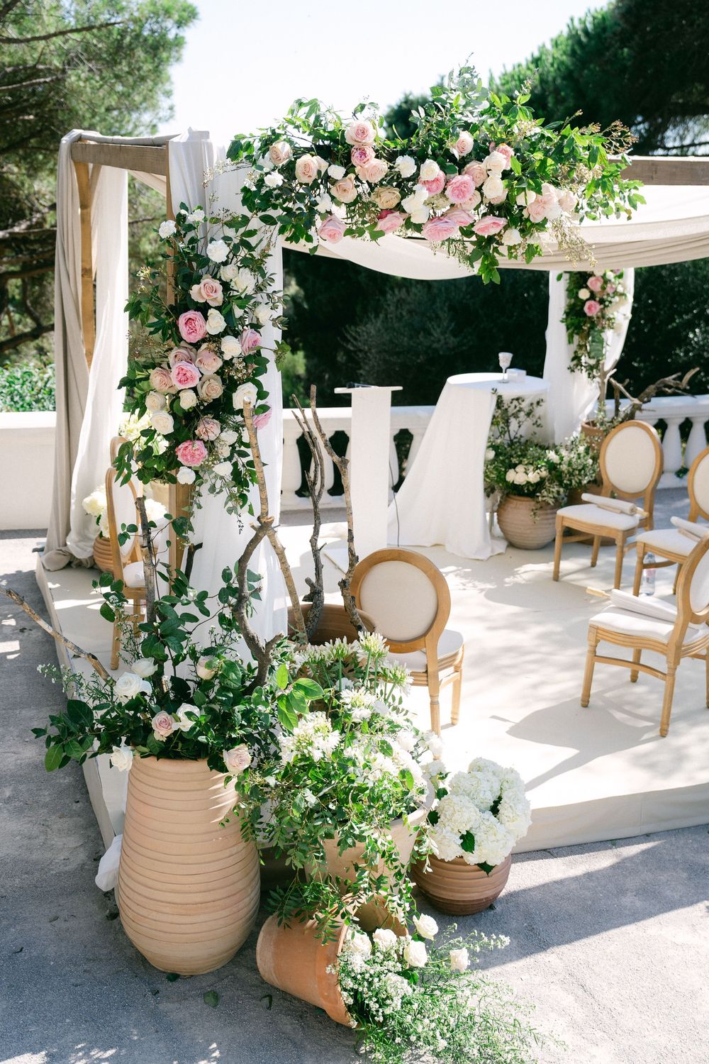 Elegant outdoor wedding arch adorned with pink roses and greenery, surrounded by terracotta pots with white floral arrangements.