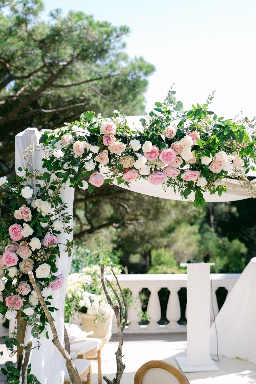 Elegant outdoor wedding arch adorned with pink and white roses and lush greenery against a sunny sky.
