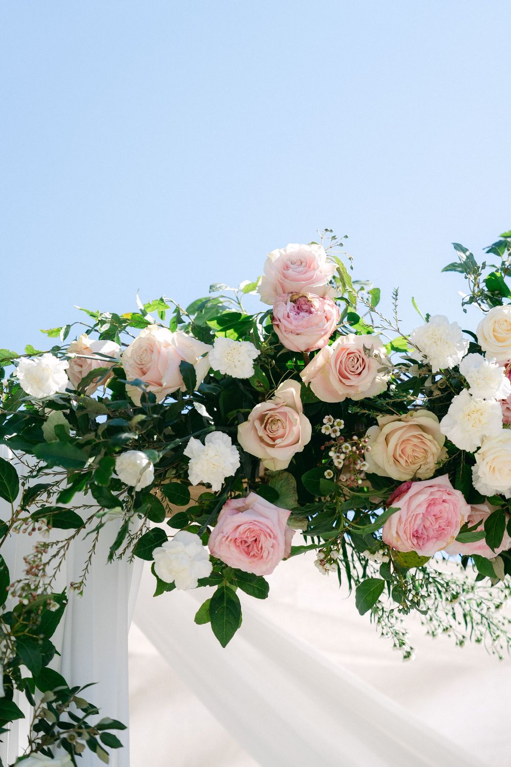 Pink and white roses with green foliage adorn a wedding arch draped in white fabric against a clear blue sky.