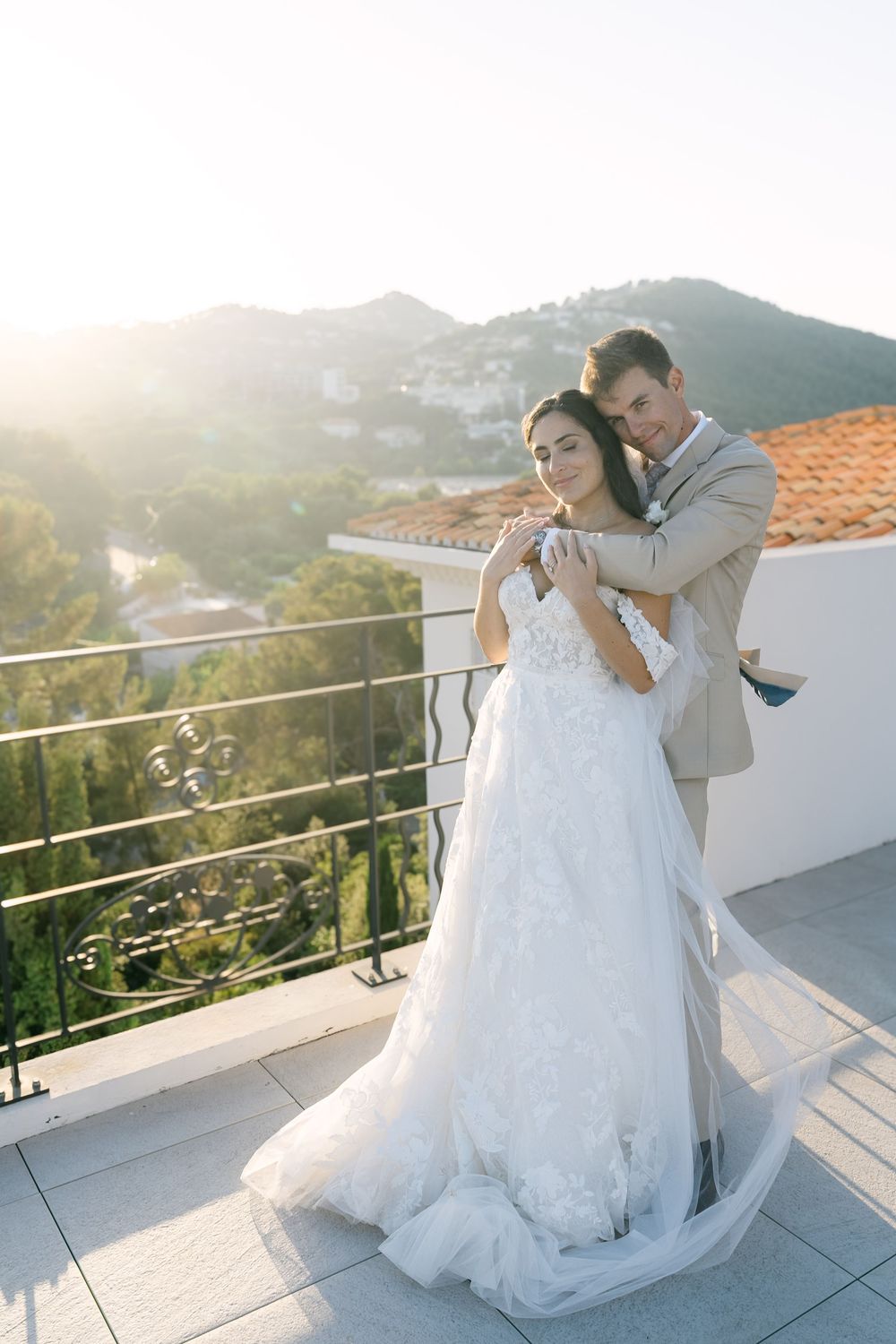 Bride and groom embracing on a sunlit balcony with scenic mountain views, bride in elegant white gown.