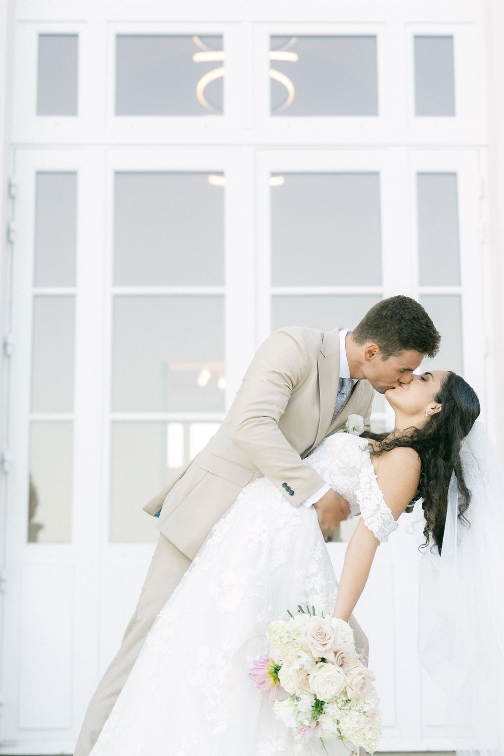 Bride and groom share a romantic dip kiss in front of white French doors, bride holding a white and pink bouquet.