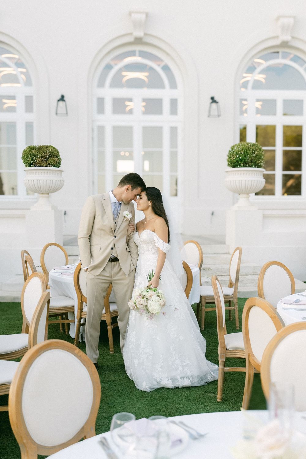 Bride and groom share a romantic kiss at an elegant outdoor wedding reception with white chairs and lush greenery.