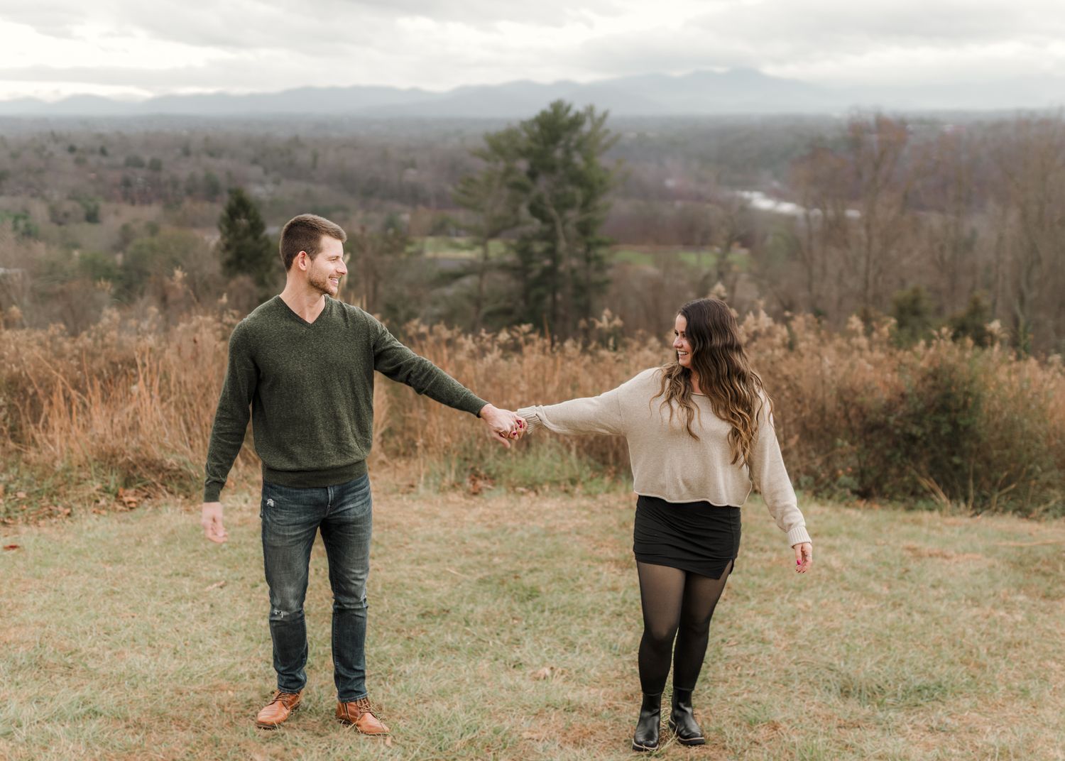 Couple holding hands in a grassy field with mountain views and autumn foliage in the background.