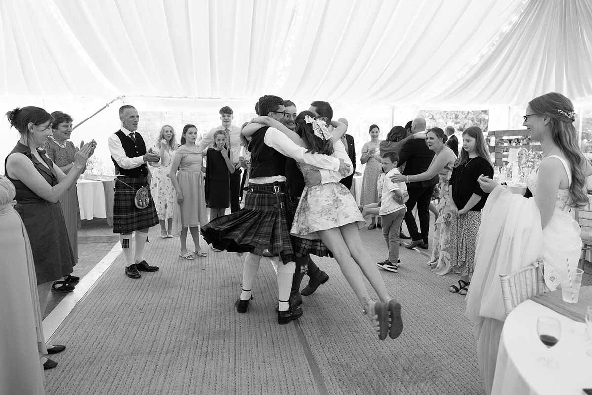 Guests dancing energetically in kilts at a Scottish wedding reception inside a marquee tent.