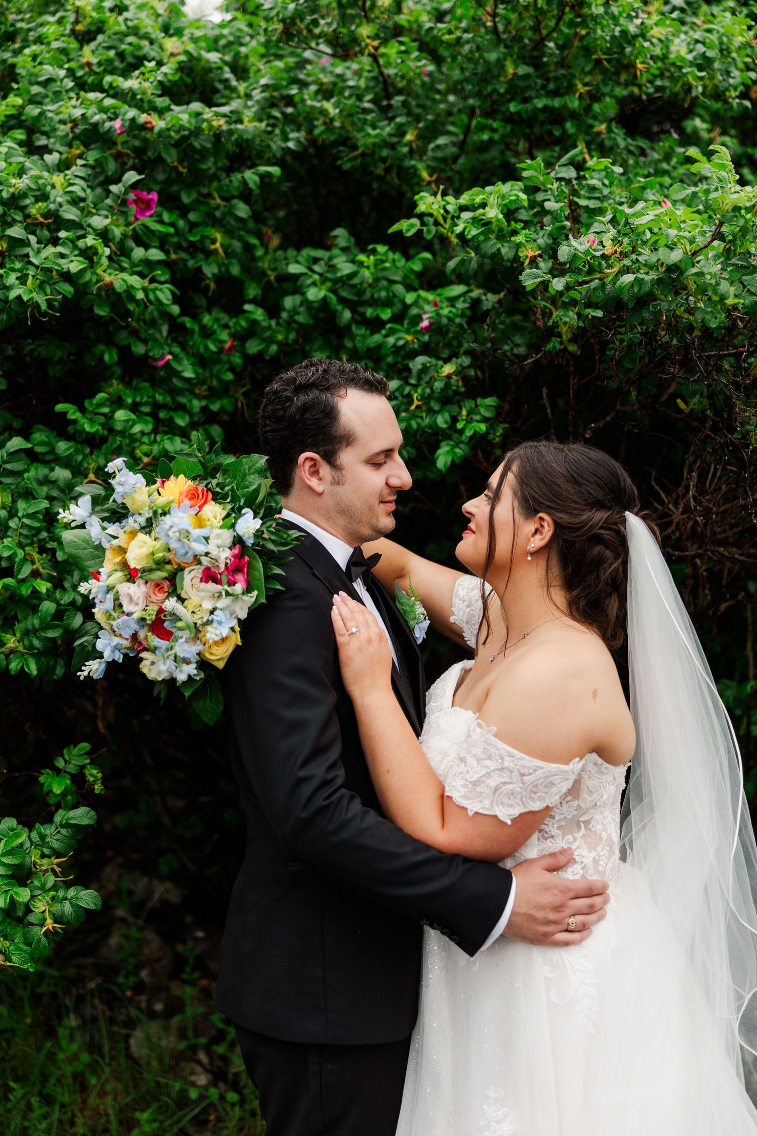 A bride and groom share a romantic moment outdoors, surrounded by lush greenery, as she holds a colorful bouquet.