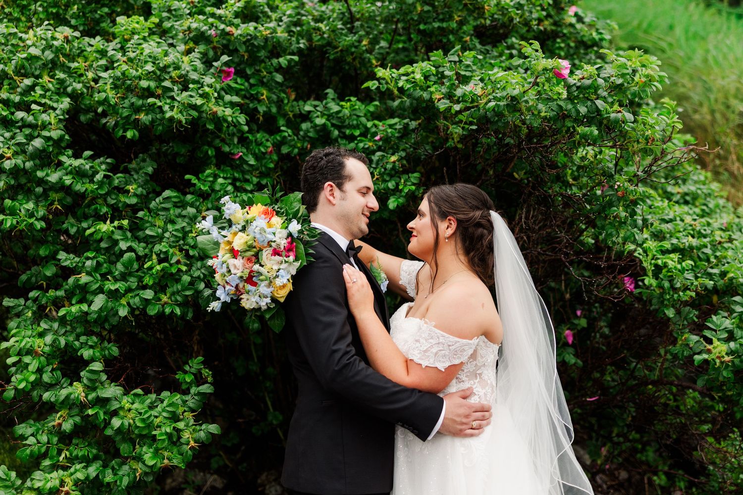 Bride and groom embrace near lush green bushes, she holds colorful bouquet, wearing white gown and veil.