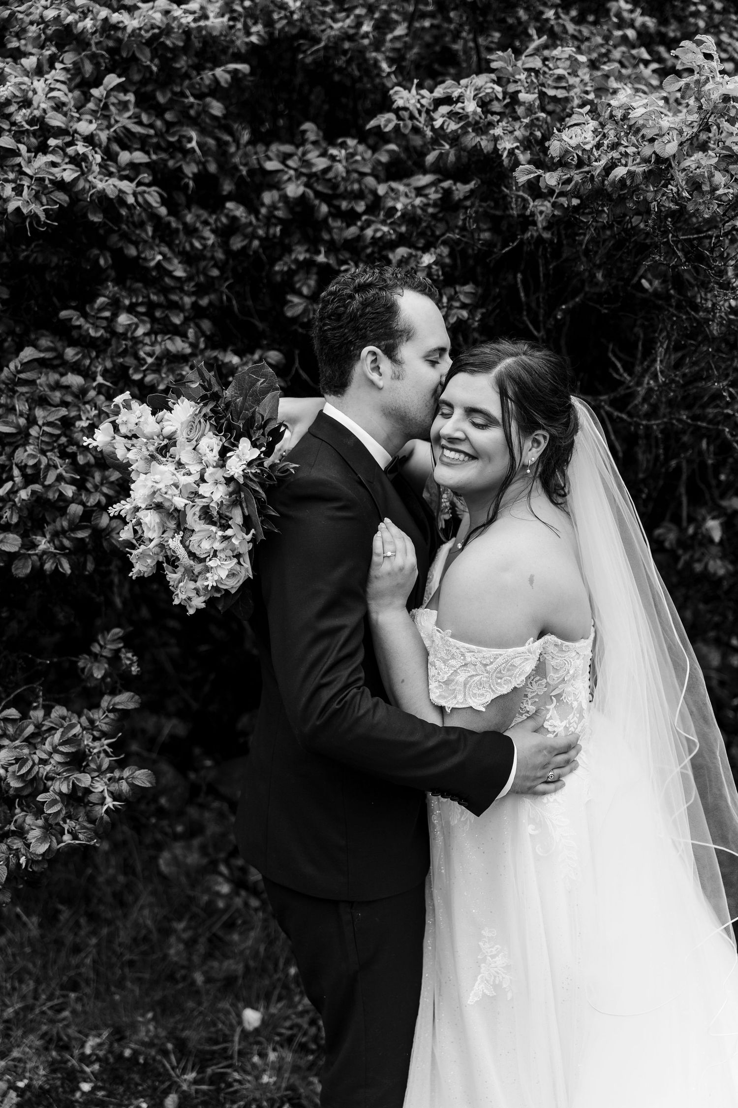 Black and white photo of a groom kissing his bride on the cheek as she smiles, holding a floral bouquet outdoors.