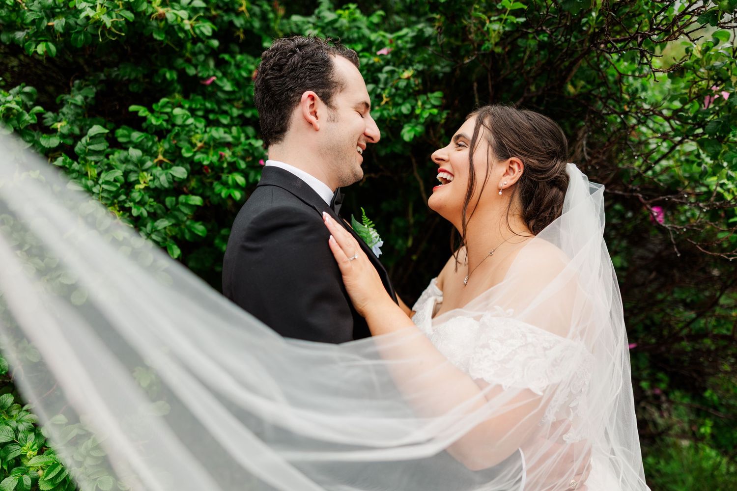 Joyful bride and groom laughing together outdoors, her veil flowing in the breeze, surrounded by lush green foliage.