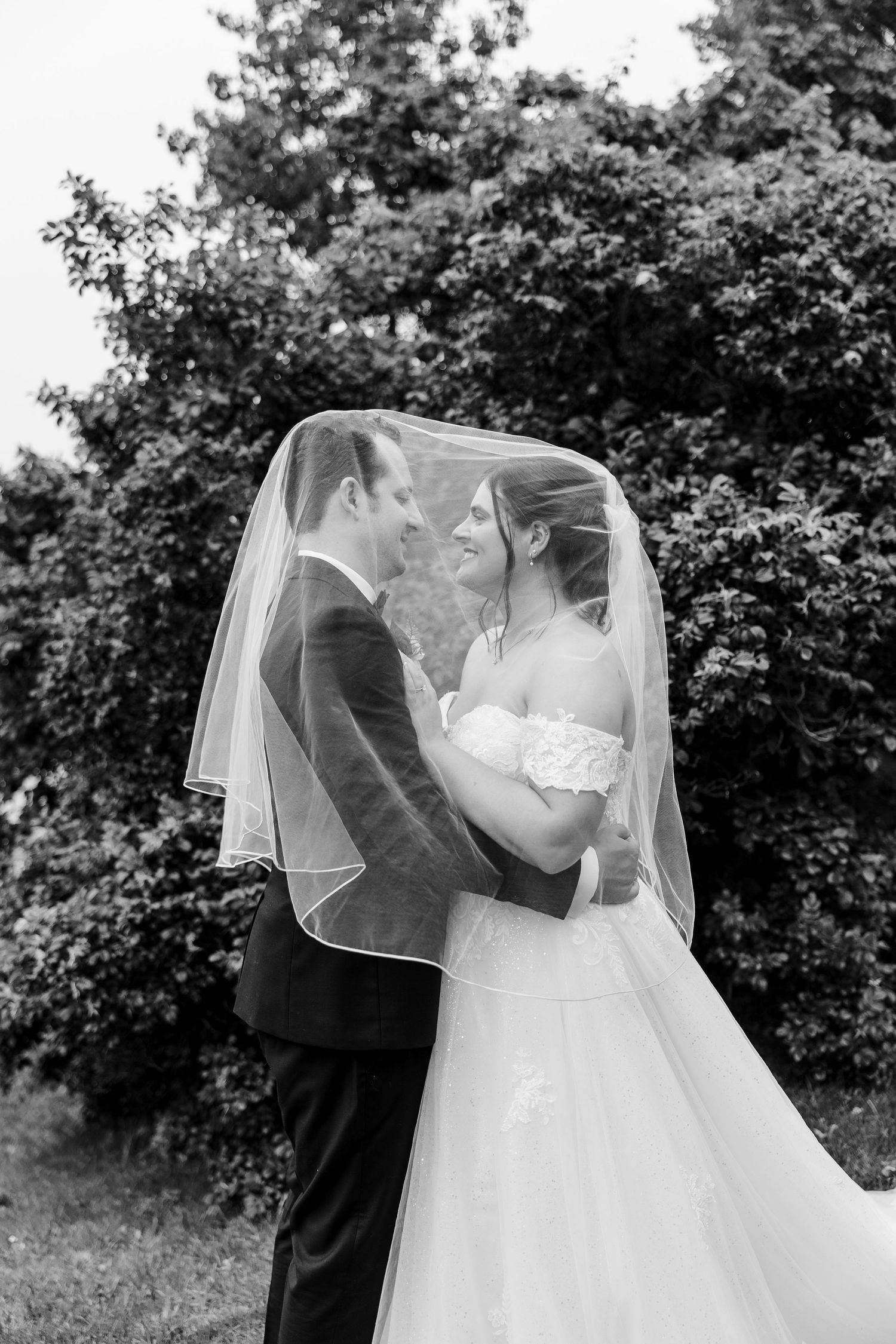 Black and white photo of bride and groom sharing a romantic moment under a wedding veil outdoors.
