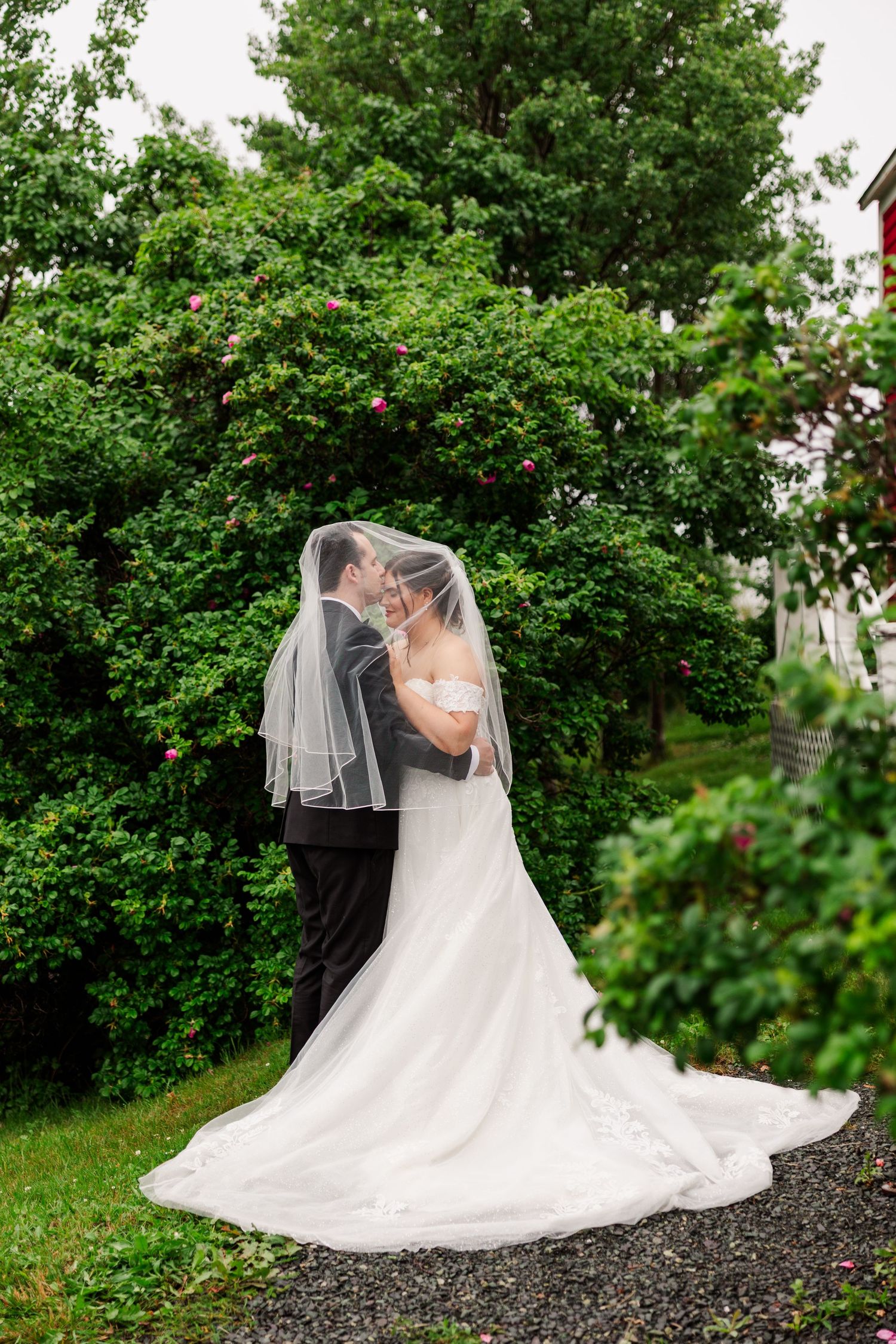 Bride and groom share romantic moment under wedding veil surrounded by lush green trees outdoors.