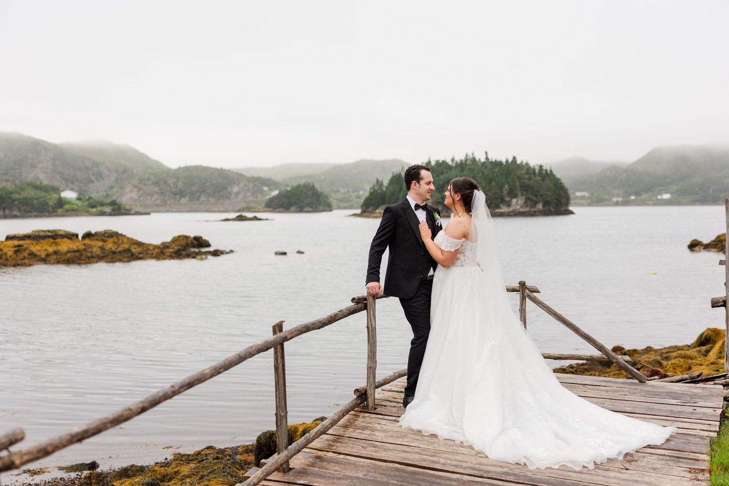 Bride and groom share a romantic moment on a rustic wooden dock overlooking a misty coastal bay on their wedding day.