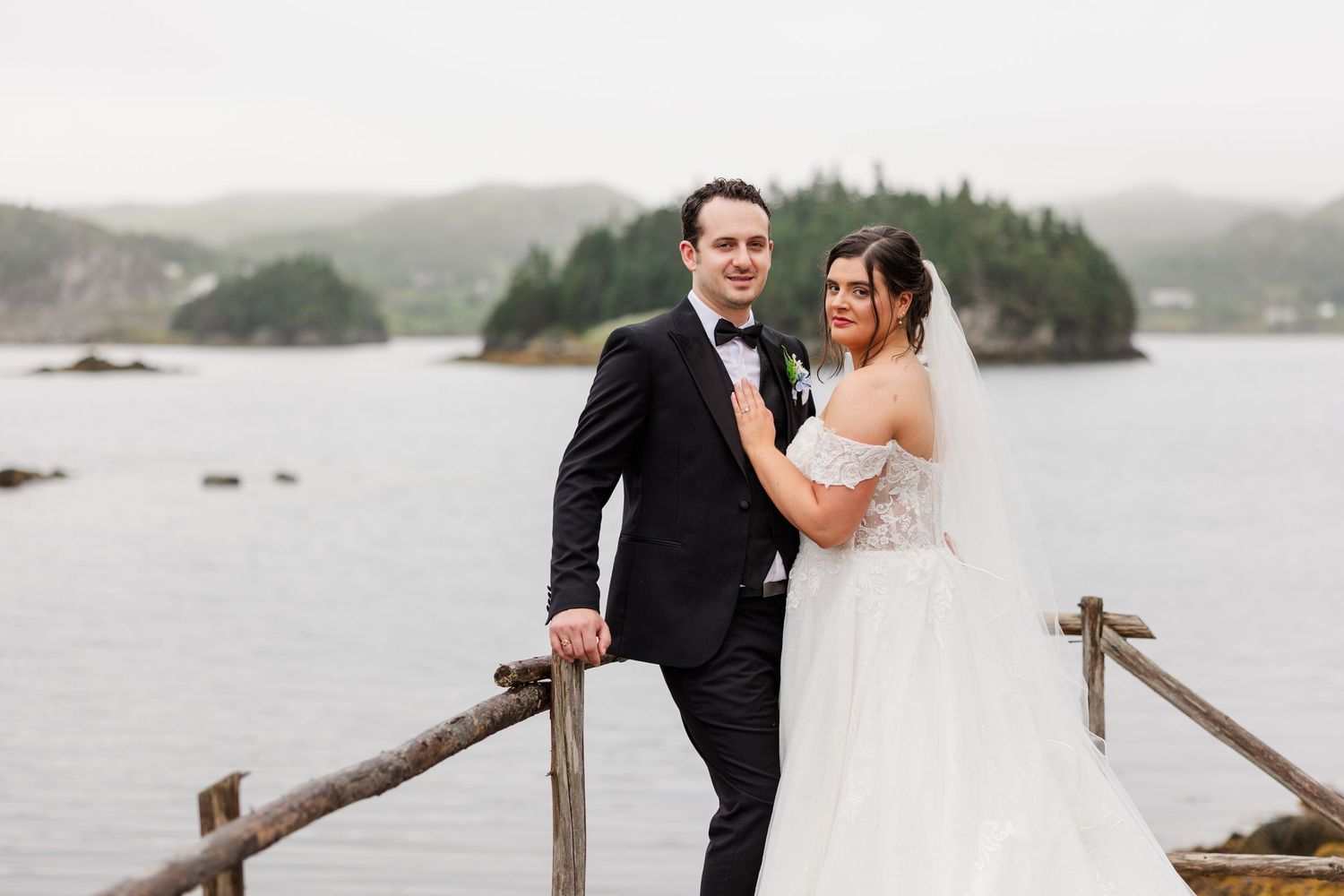 Bride and groom pose on a waterfront dock with misty ocean and island backdrop on their wedding day.